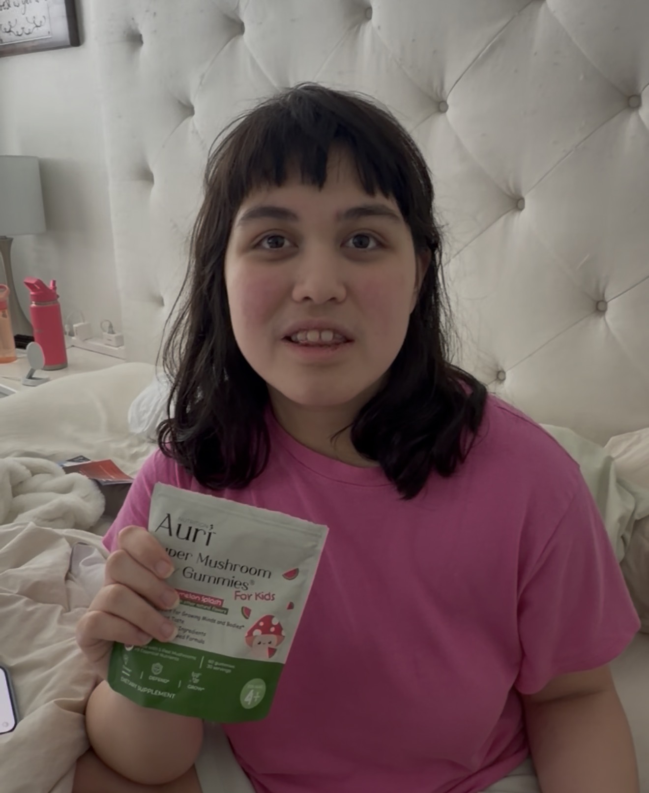 Young woman with dark hair and bangs holding a pamphlet, wearing a pink shirt, sitting on a bed with a tufted headboard.