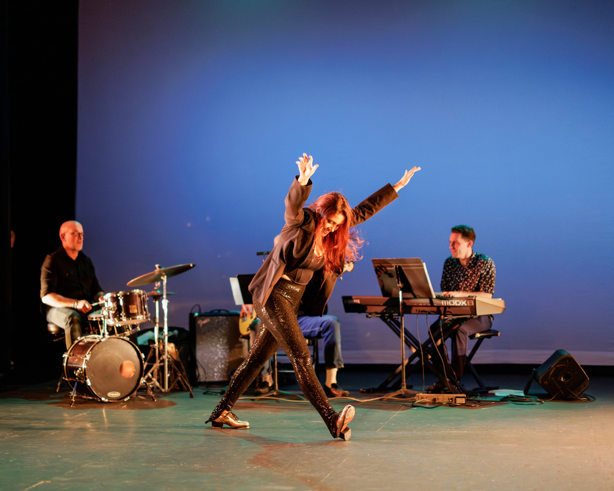 Two dancers perform on stage with musicians playing instruments in the background, against a blue backdrop.