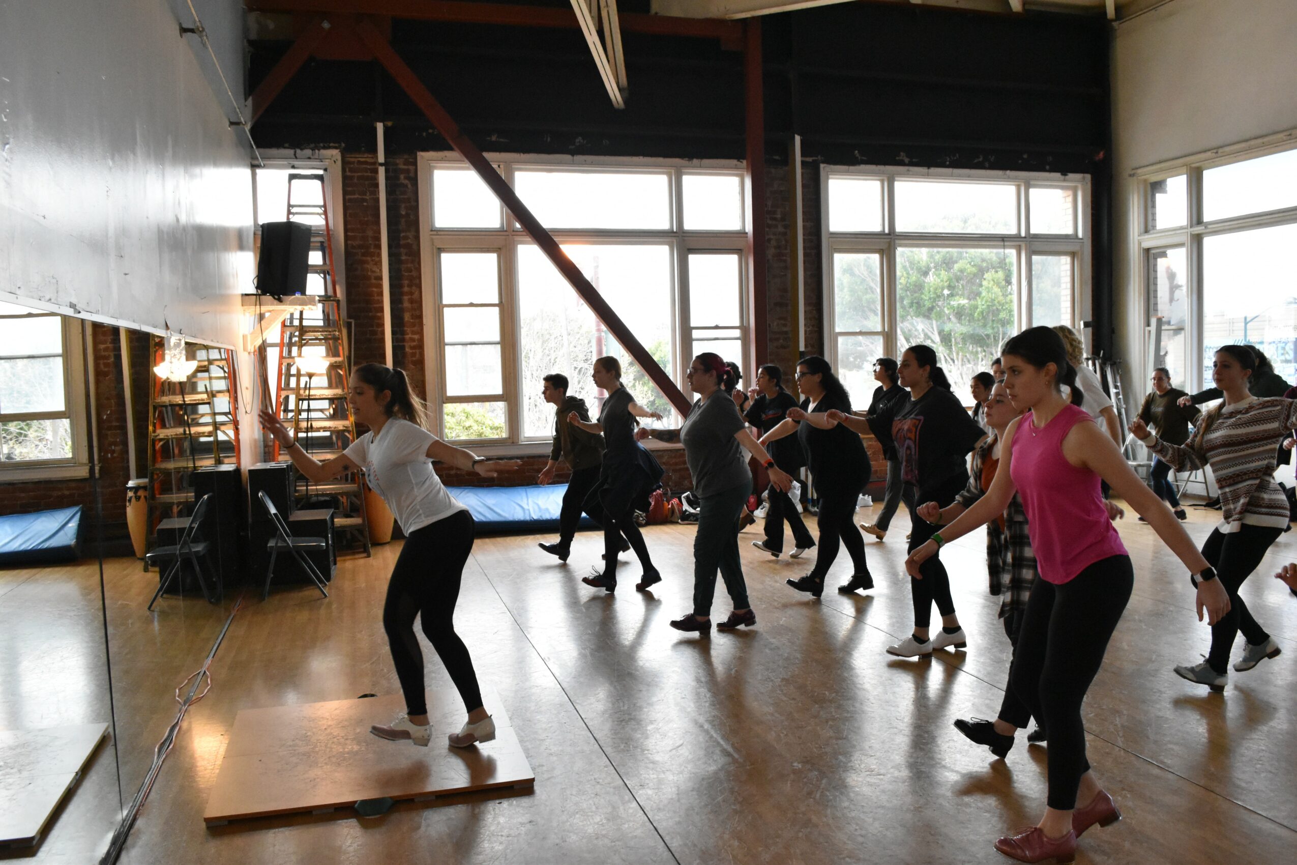 Group of people participating in a dance or exercise class in a spacious room with large windows, wooden floors, and natural light.
