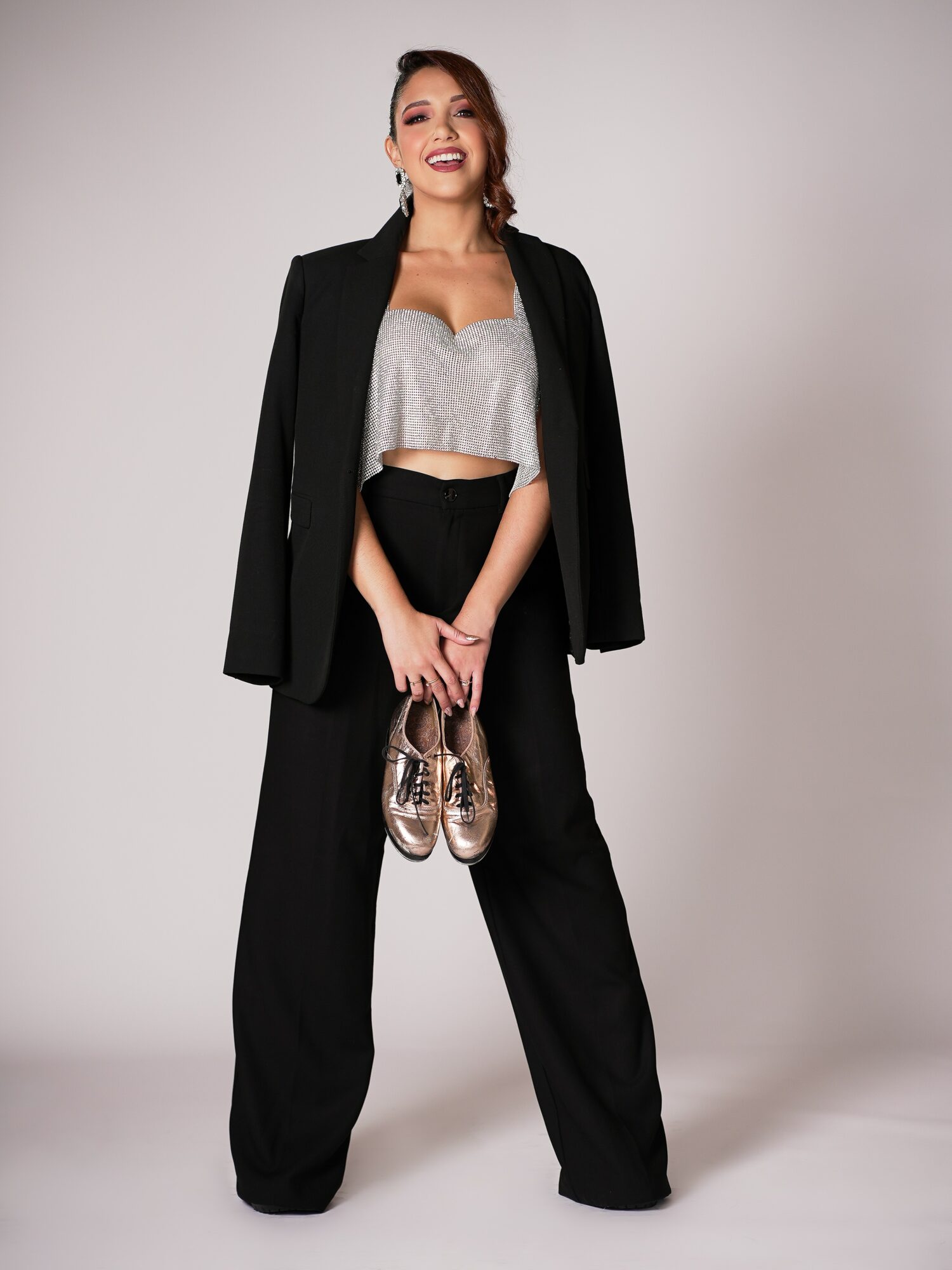 Young woman with long hair in a ponytail, smiling, wearing a black blazer, silver crop top, black wide-leg pants, holding sneakers.