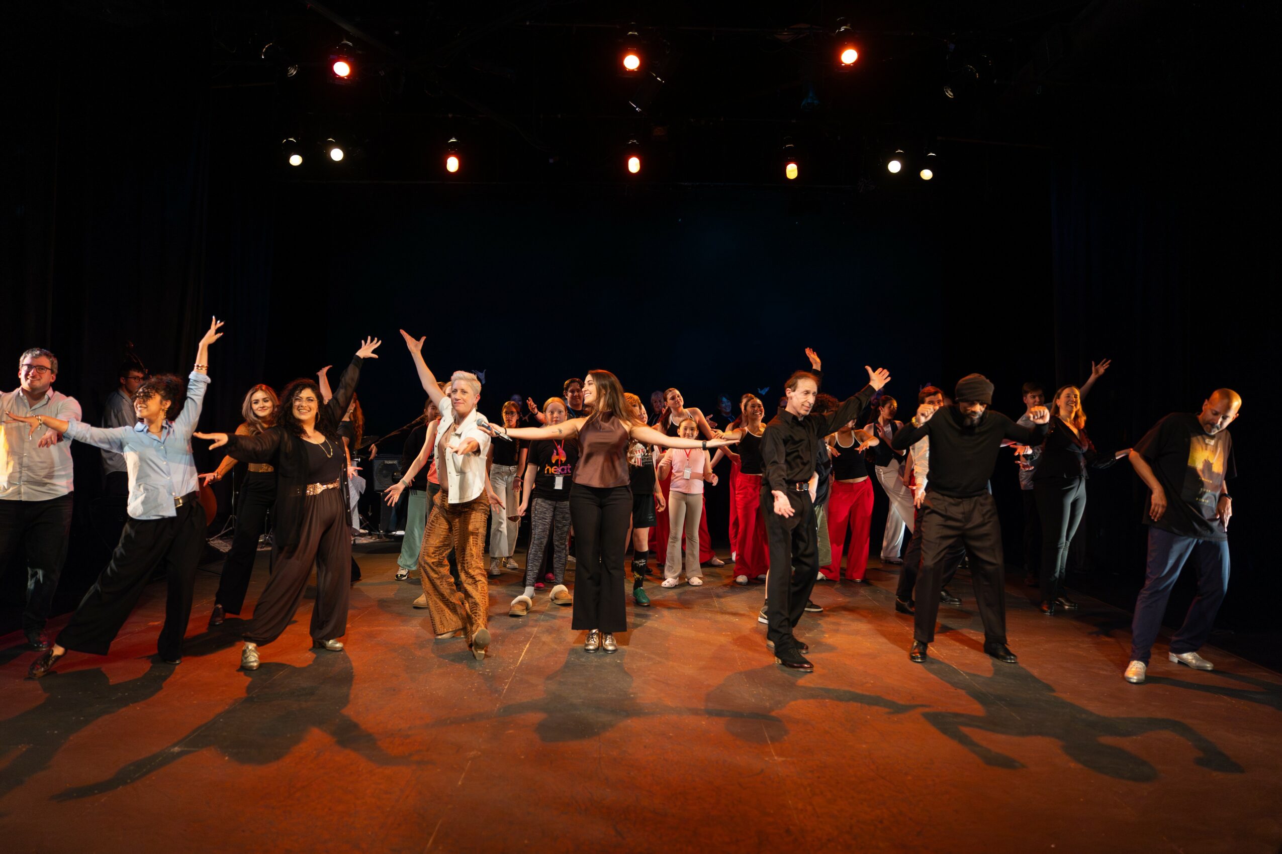 Group of people dancing on stage with stage lights overhead, some raising arms, in a performance setting.