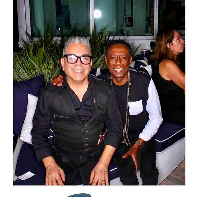 Two men sitting together smiling, one with glasses and gray hair, the other with dark hair, in a social setting.