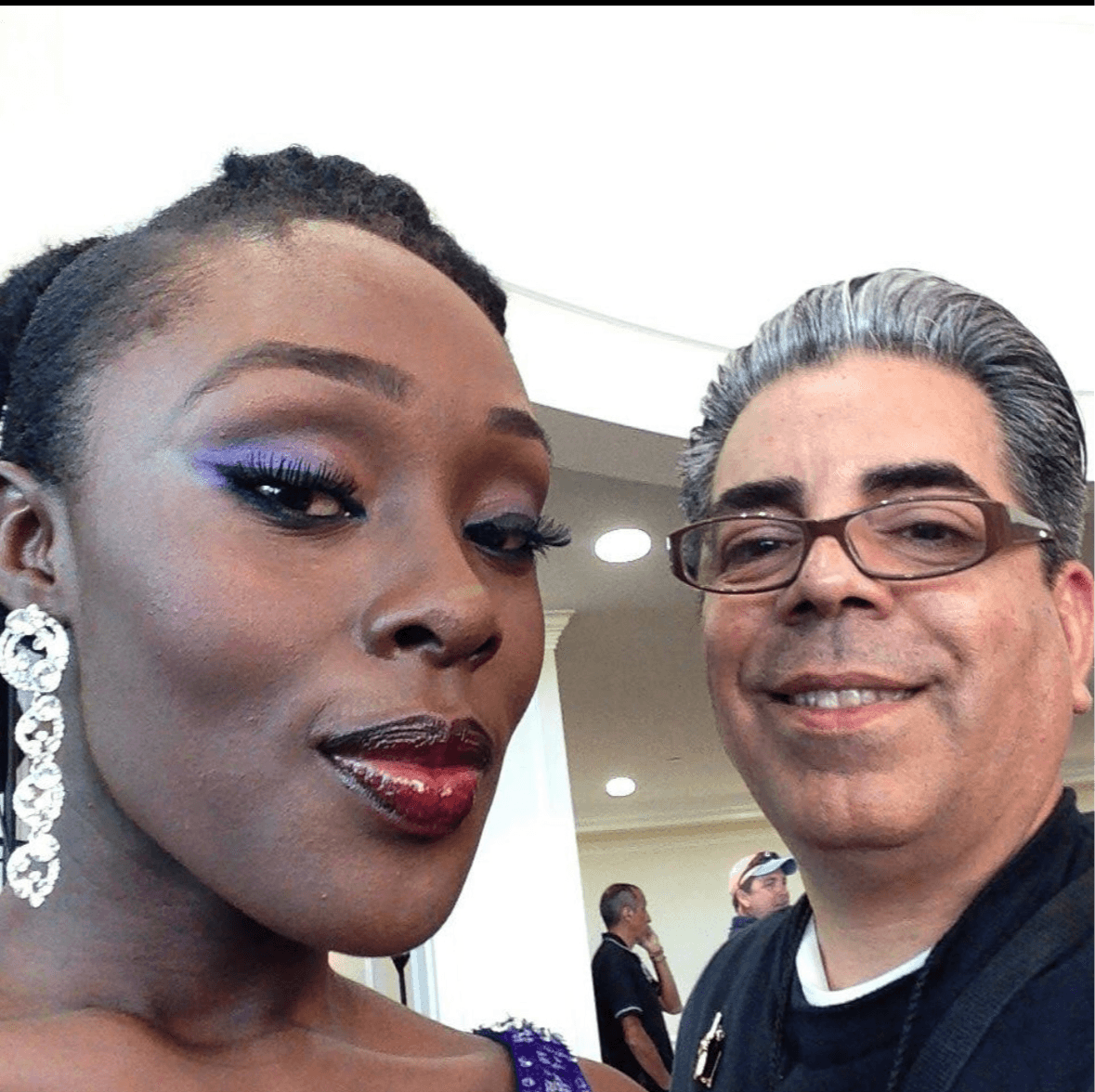 Close-up of a woman with makeup and earrings, smiling with a man wearing glasses in an indoor setting.