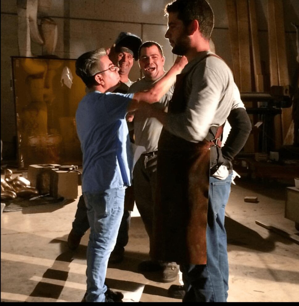 Group of five people, including a young boy, in a workshop or studio, engaging in a conversation or activity, with tools and equipment in the background.