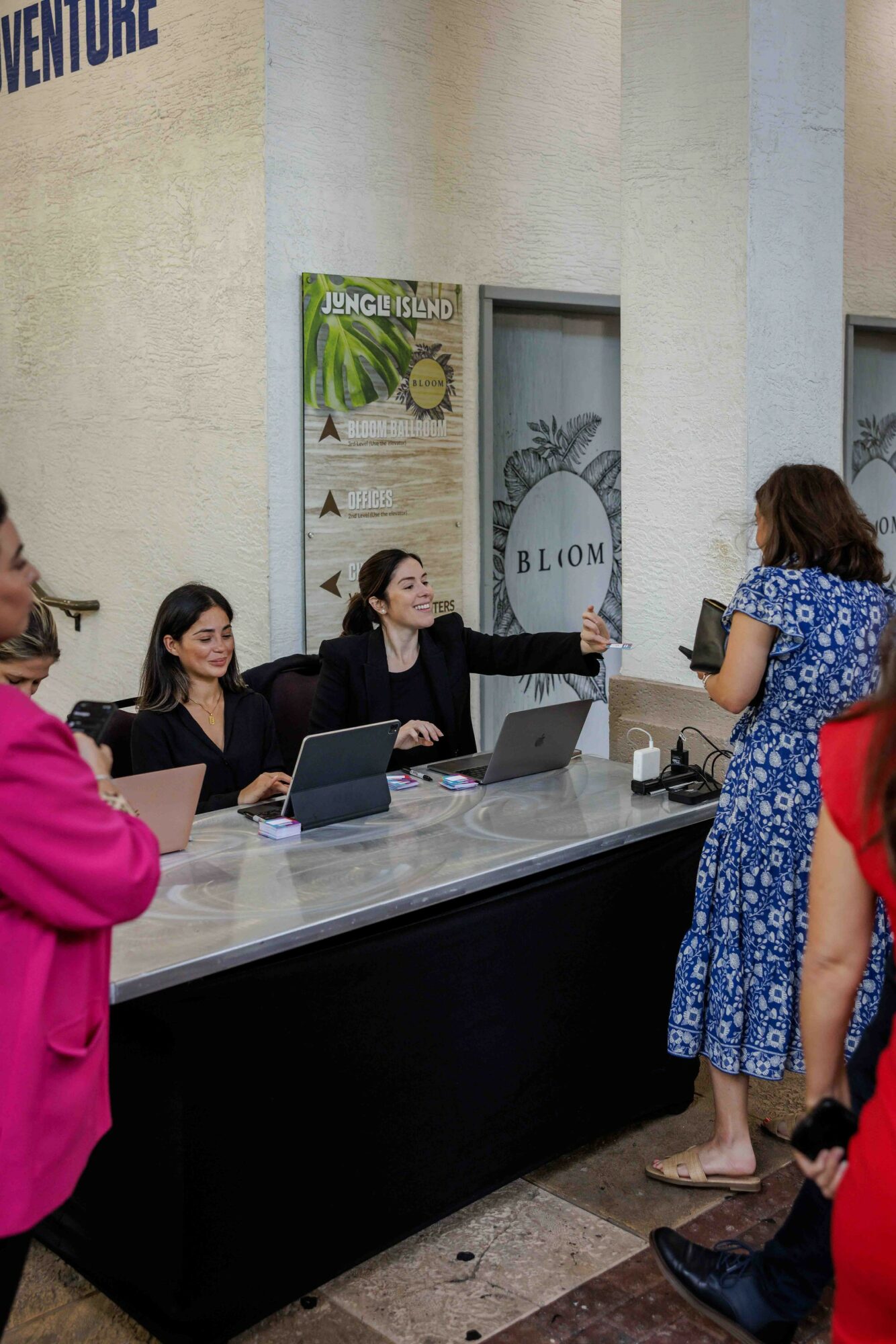 Four women sit at a table with laptops, engaging with a standing woman in a blue floral dress, in front of a wall with signs.