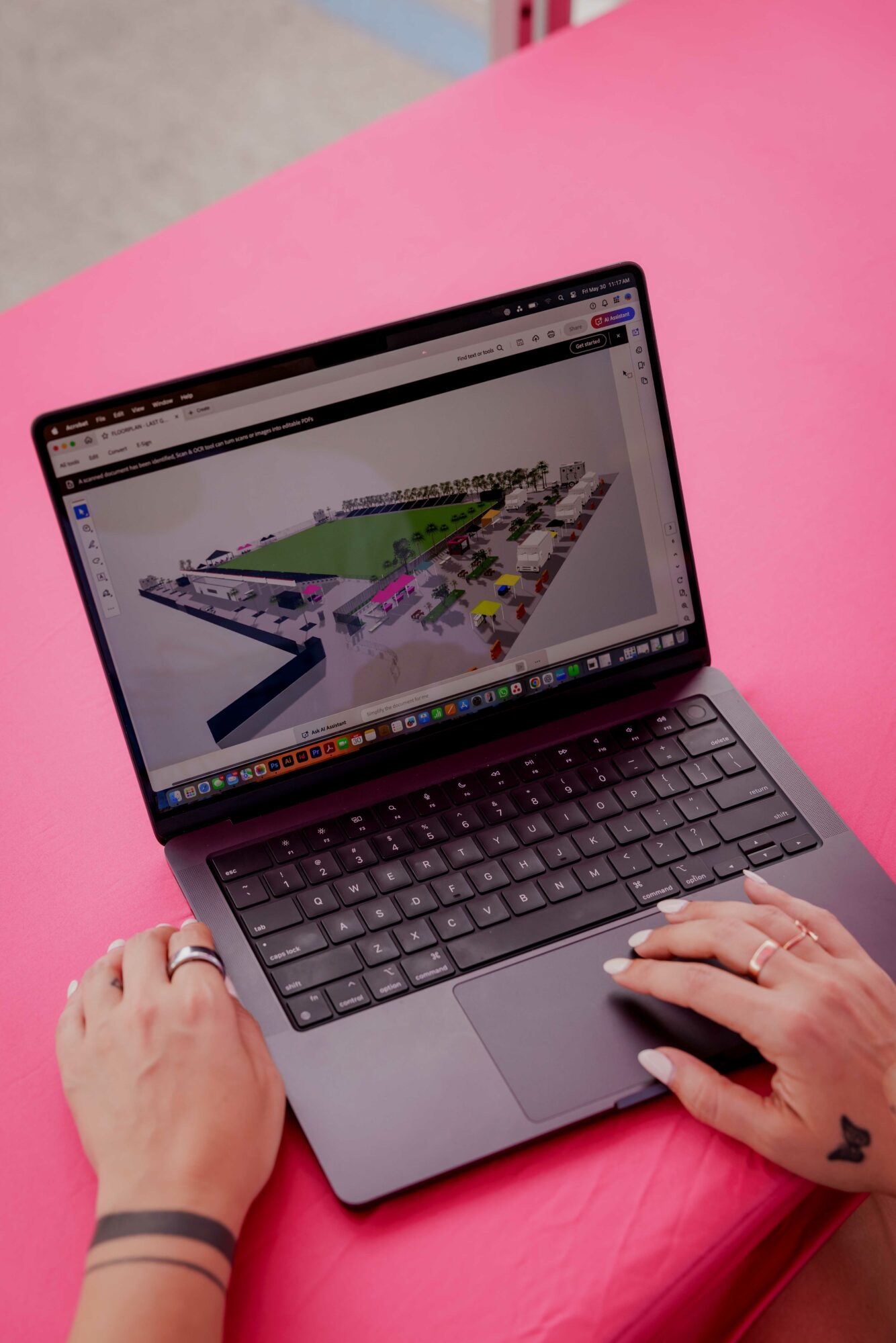 Person's hands using a laptop on a pink surface, displaying a 3D model or map on the screen.