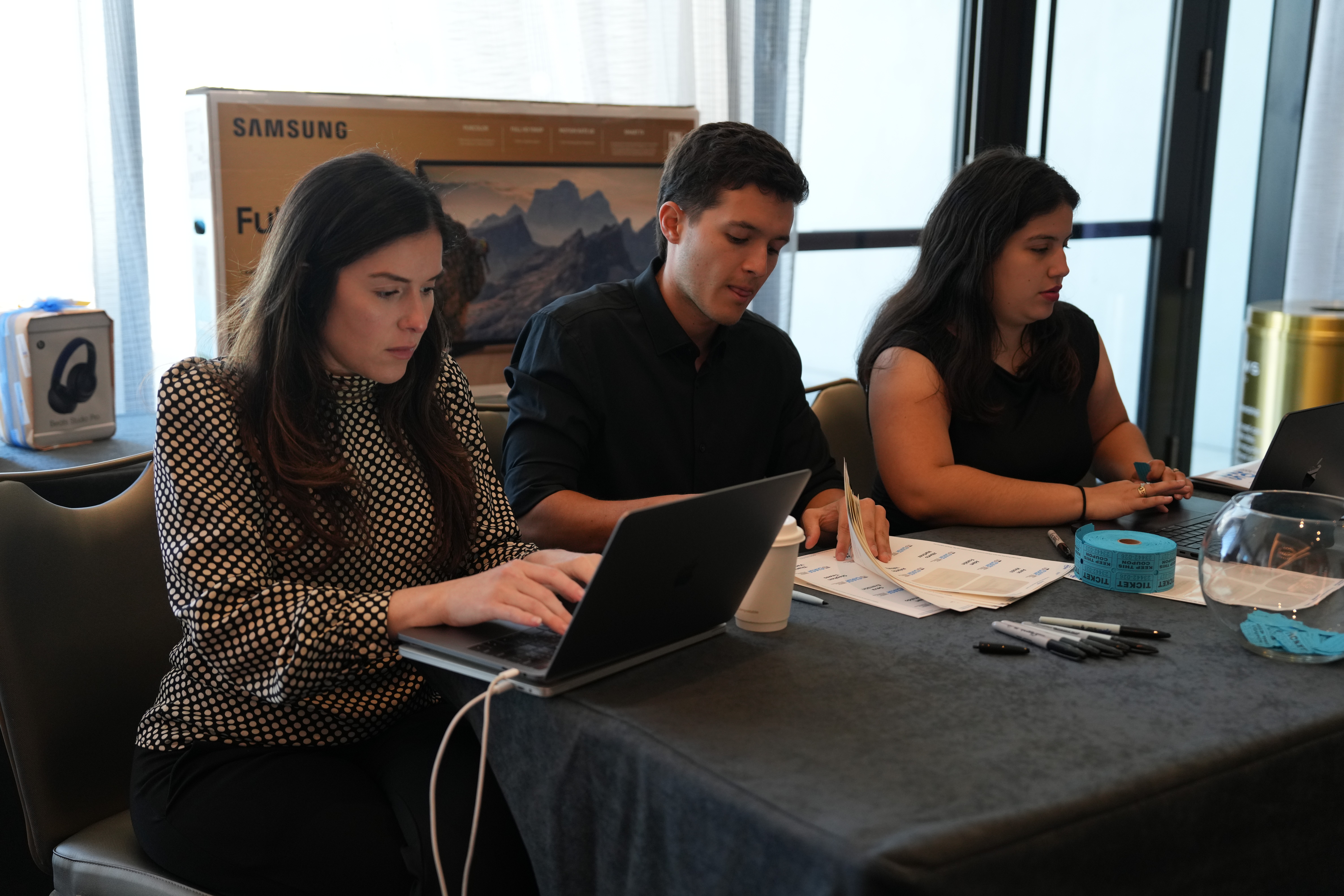 Three people sit at a table working on laptops, with a large screen and window behind them.