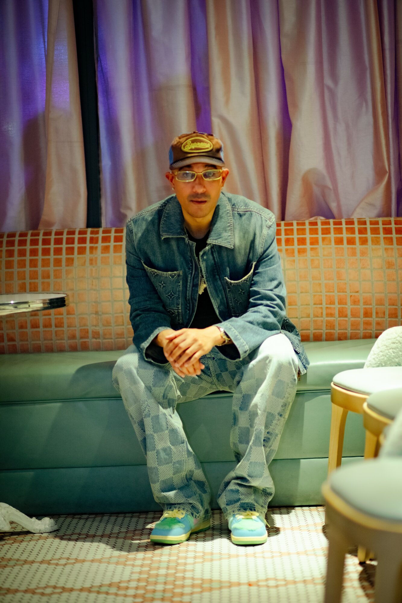 Young man sitting on a sofa with curtains in the background, wearing a denim jacket, patterned pants, and sneakers.