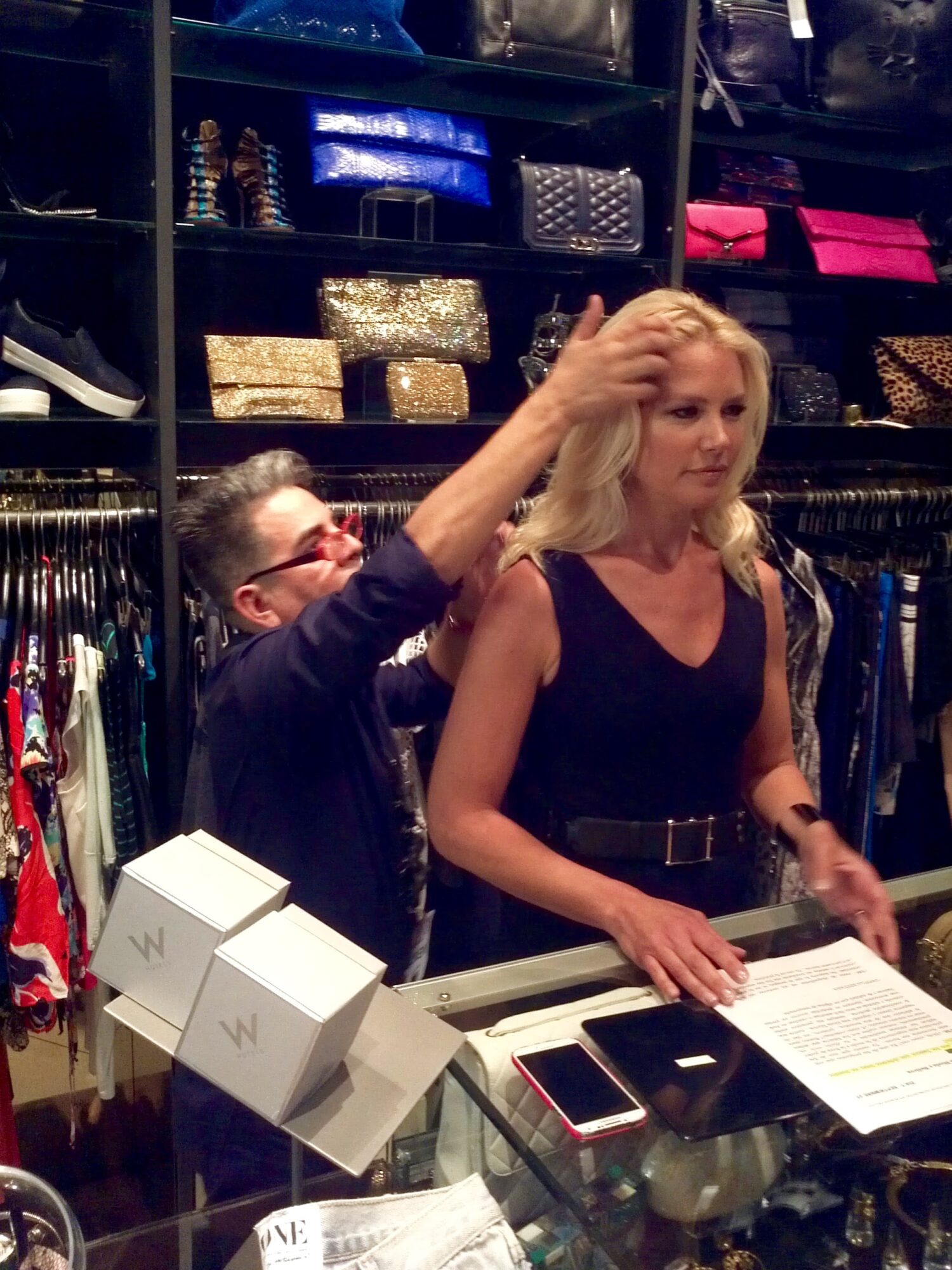 A person adjusts a woman's hair in a clothing store, with shelves of bags and clothes in the background.