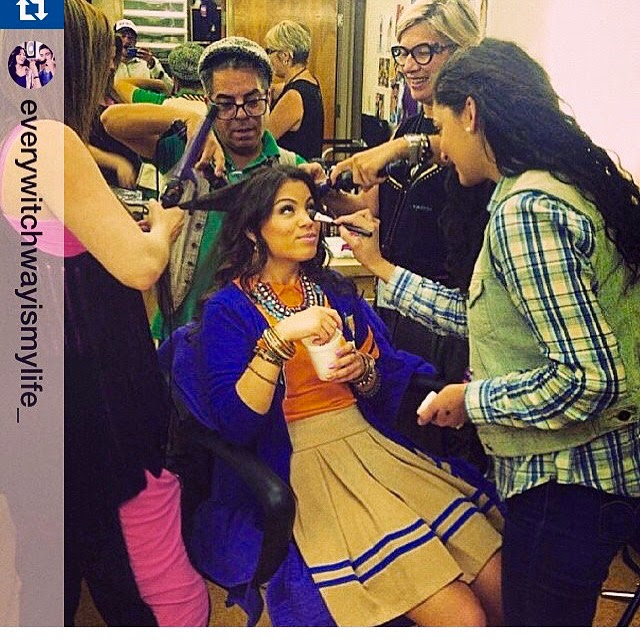 Young woman in traditional dress is surrounded by makeup artists and stylists in a busy room.