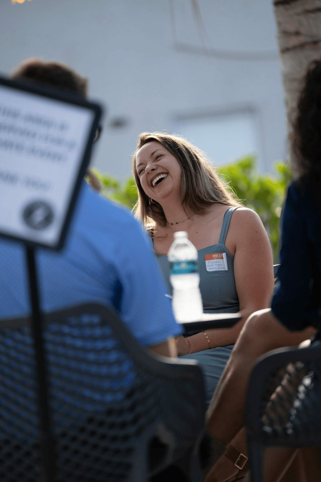 Woman laughing and talking outdoors with people, a water bottle on table, bright background.