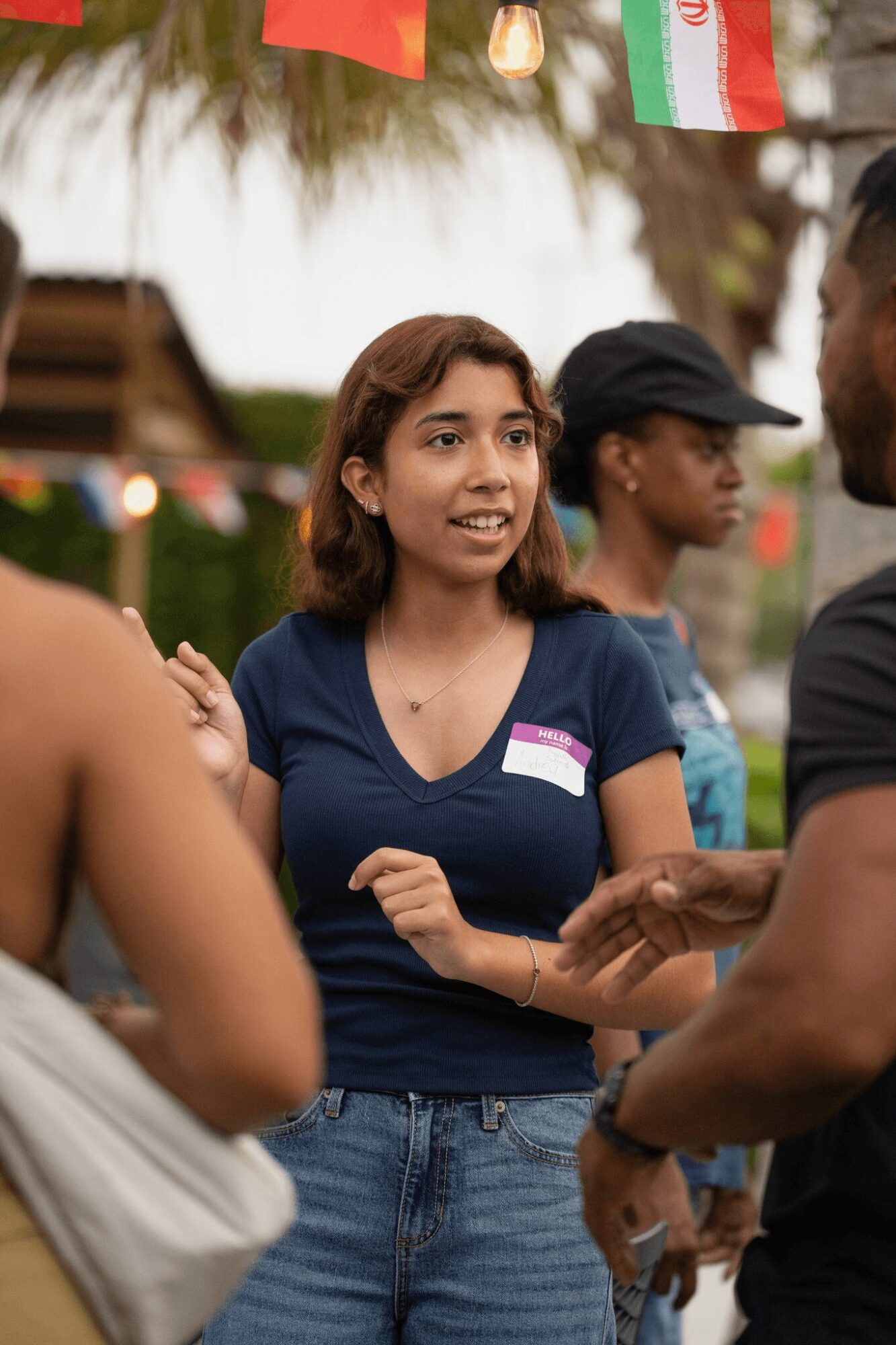 Young woman with shoulder-length hair smiling, wearing a navy blue shirt, engaging in conversation outdoors with others.