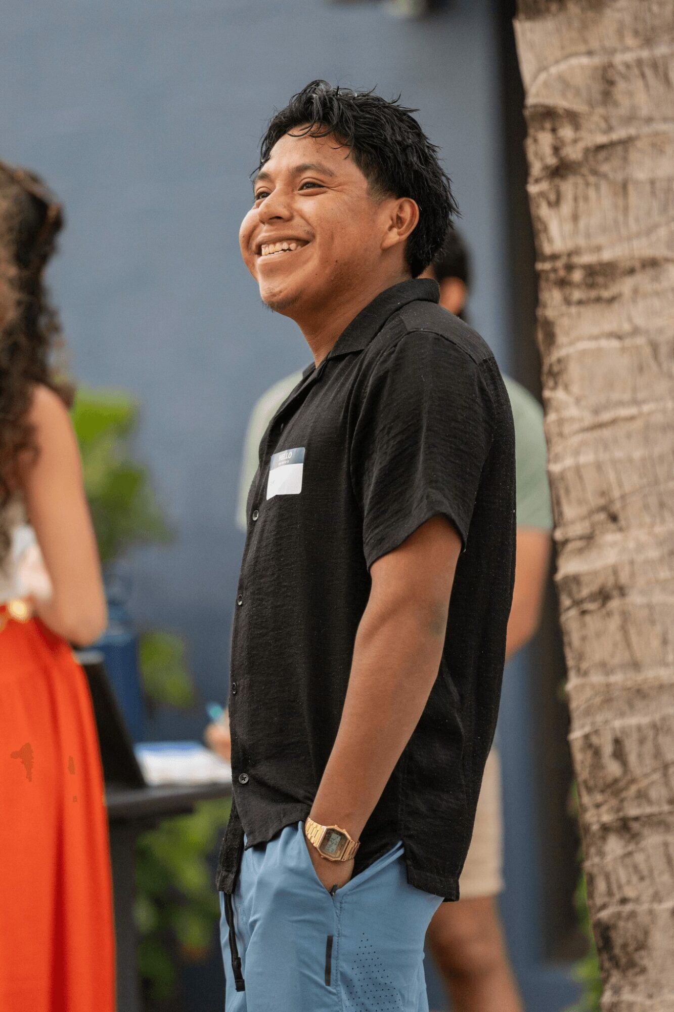 Young man smiling outdoors, wearing a black shirt and blue pants, standing near a tree.