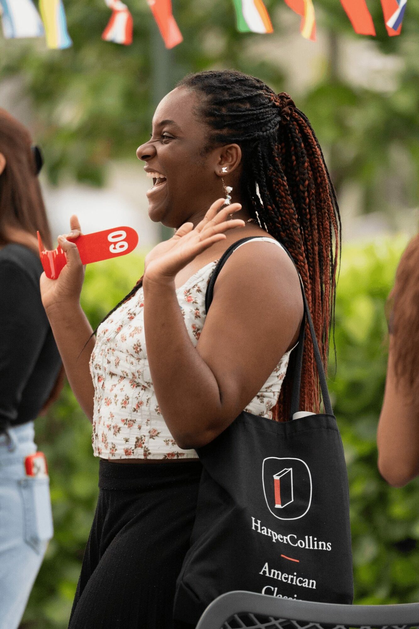Smiling woman with braided hair talking outdoors, holding a red object, with trees and people in background.