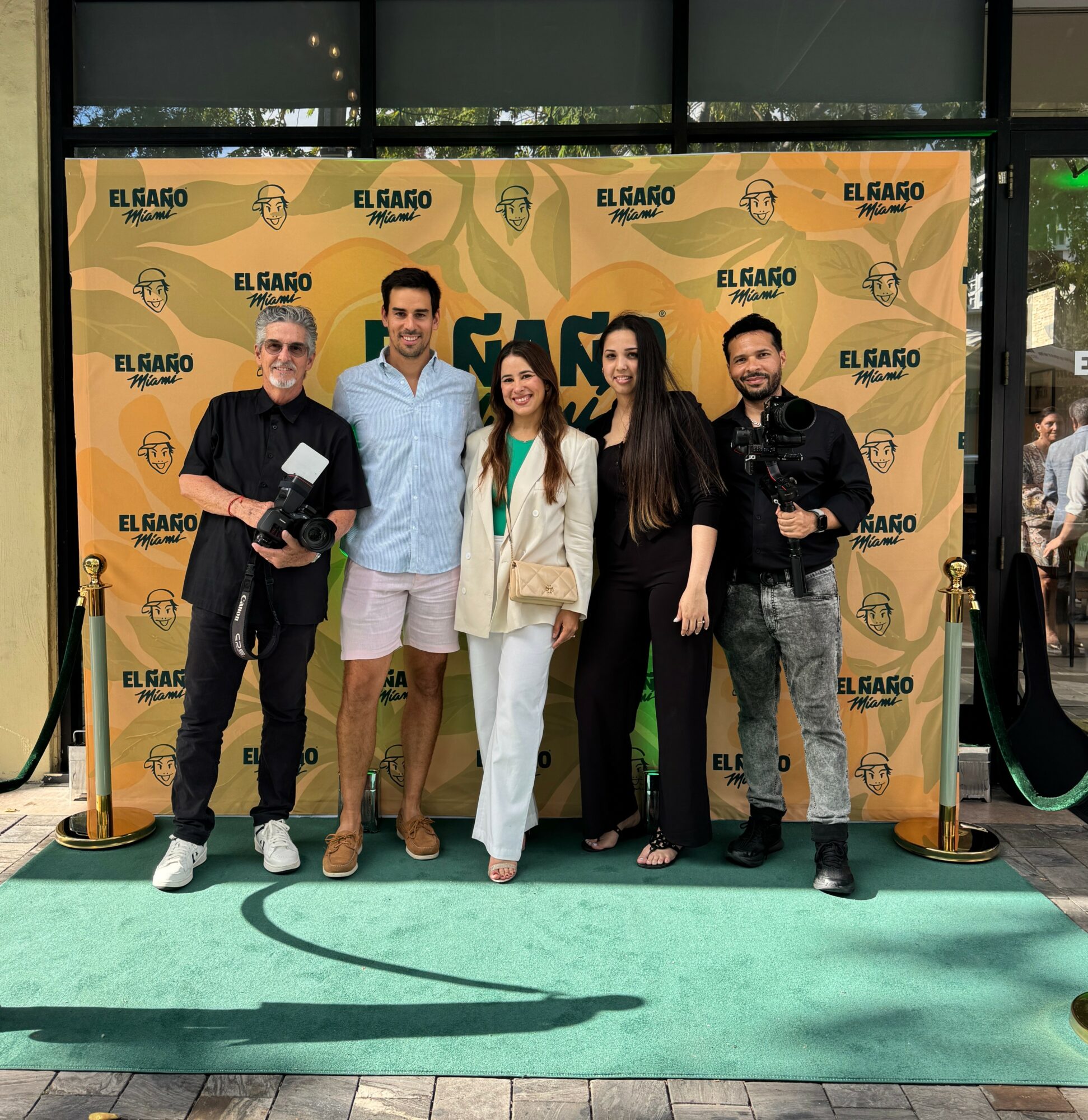 Five people standing on a green carpet in front of a yellow backdrop with logos, smiling for the photo.