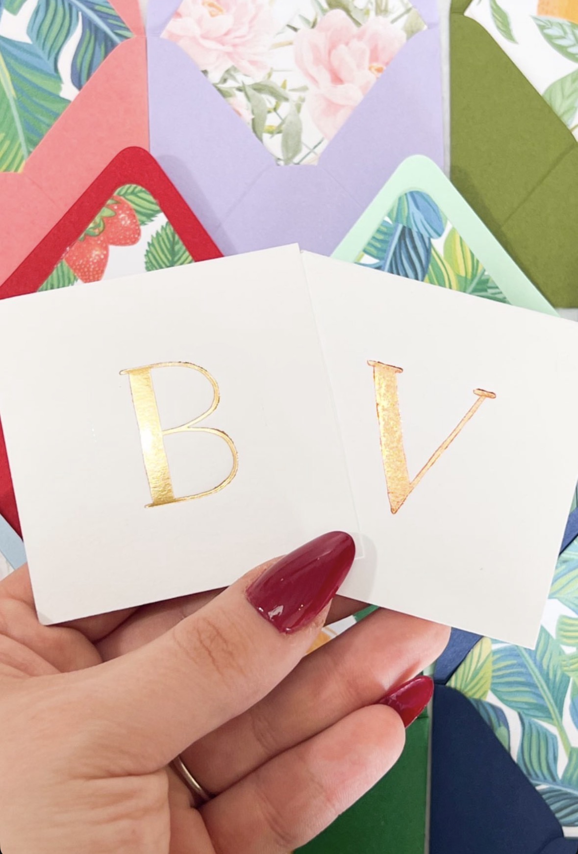 Two cards with gold letters 'B' and 'V' held by a person with red nail polish, surrounded by colorful envelopes and floral patterns.