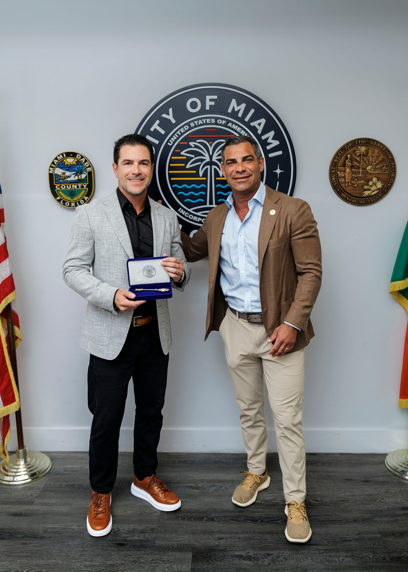 Two men standing in front of a Miami city emblem, holding an award, with flags on either side.