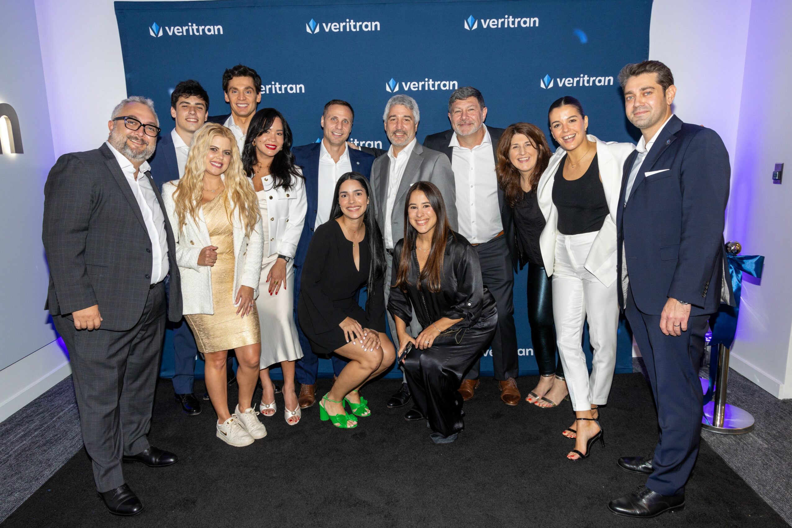 Group of 13 people posing in front of a blue Veritron backdrop, some standing and some sitting, in a professional setting.