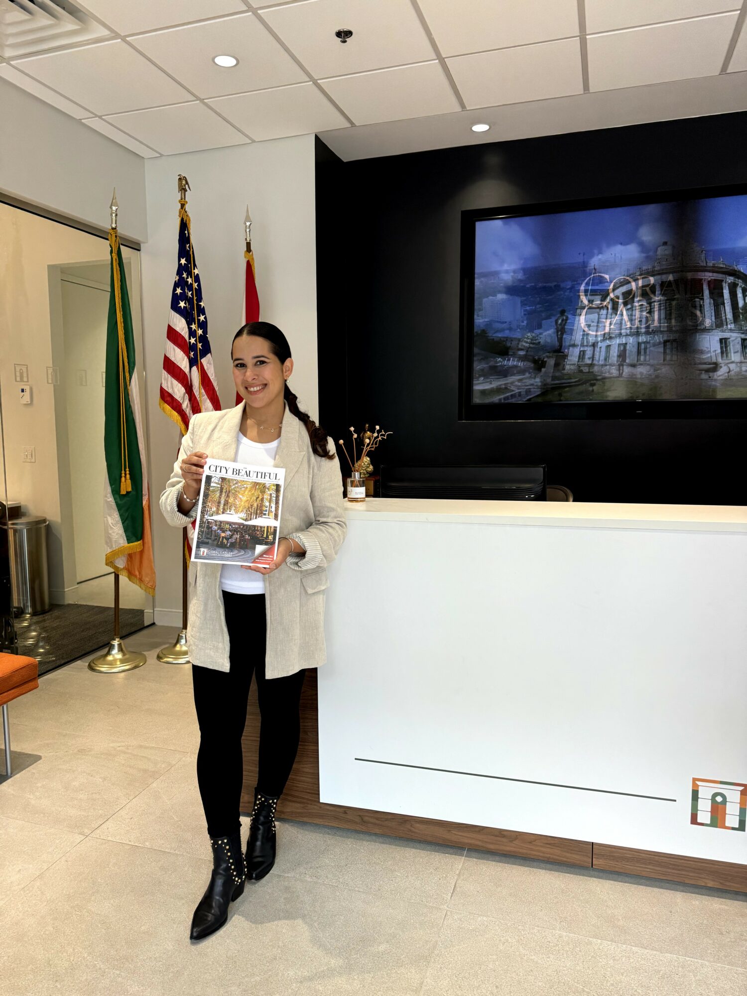 Woman standing indoors holding a magazine, smiling, with flags and a large screen behind her.