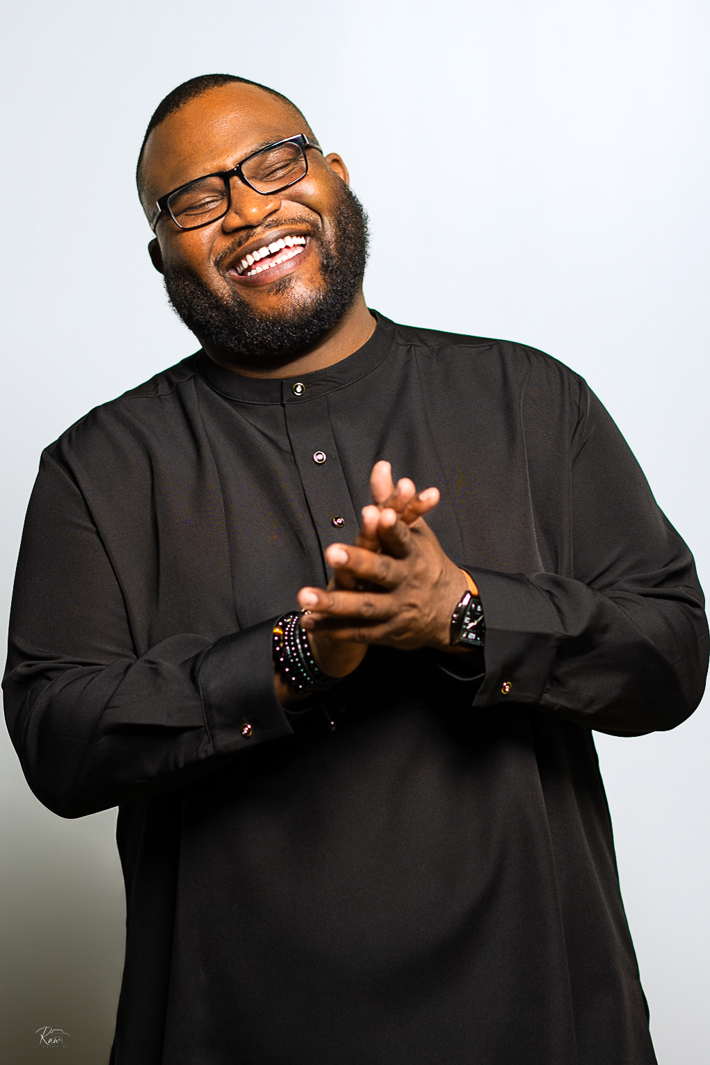 Smiling man with glasses and a beard, wearing a black shirt, clapping hands, against a light background.