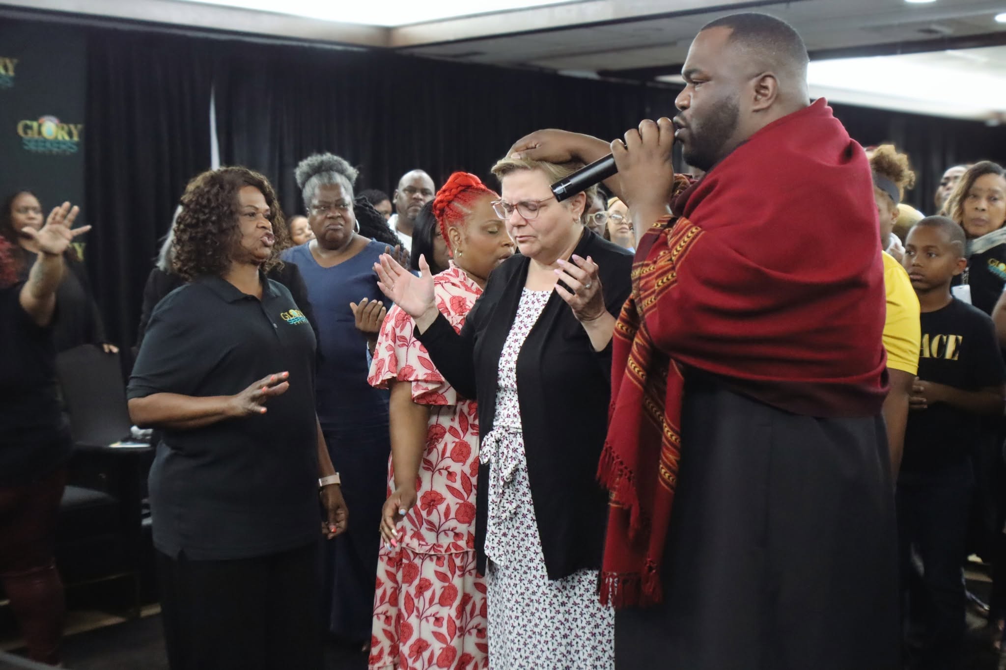 Group of people gathered indoors, some with hands raised, listening to a man speaking into a microphone, diverse ages and styles.