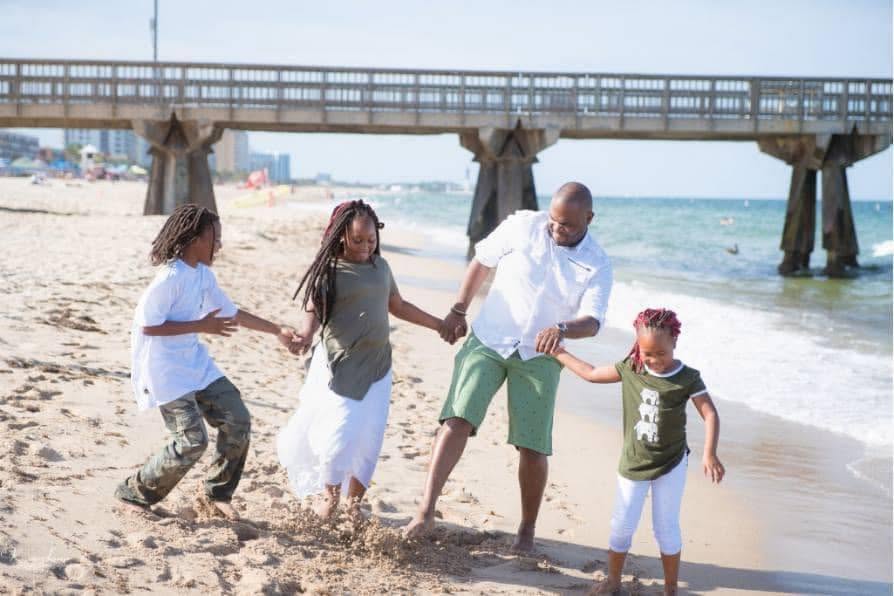 Family of four holding hands and walking on the beach near a pier, with ocean and sky in background.