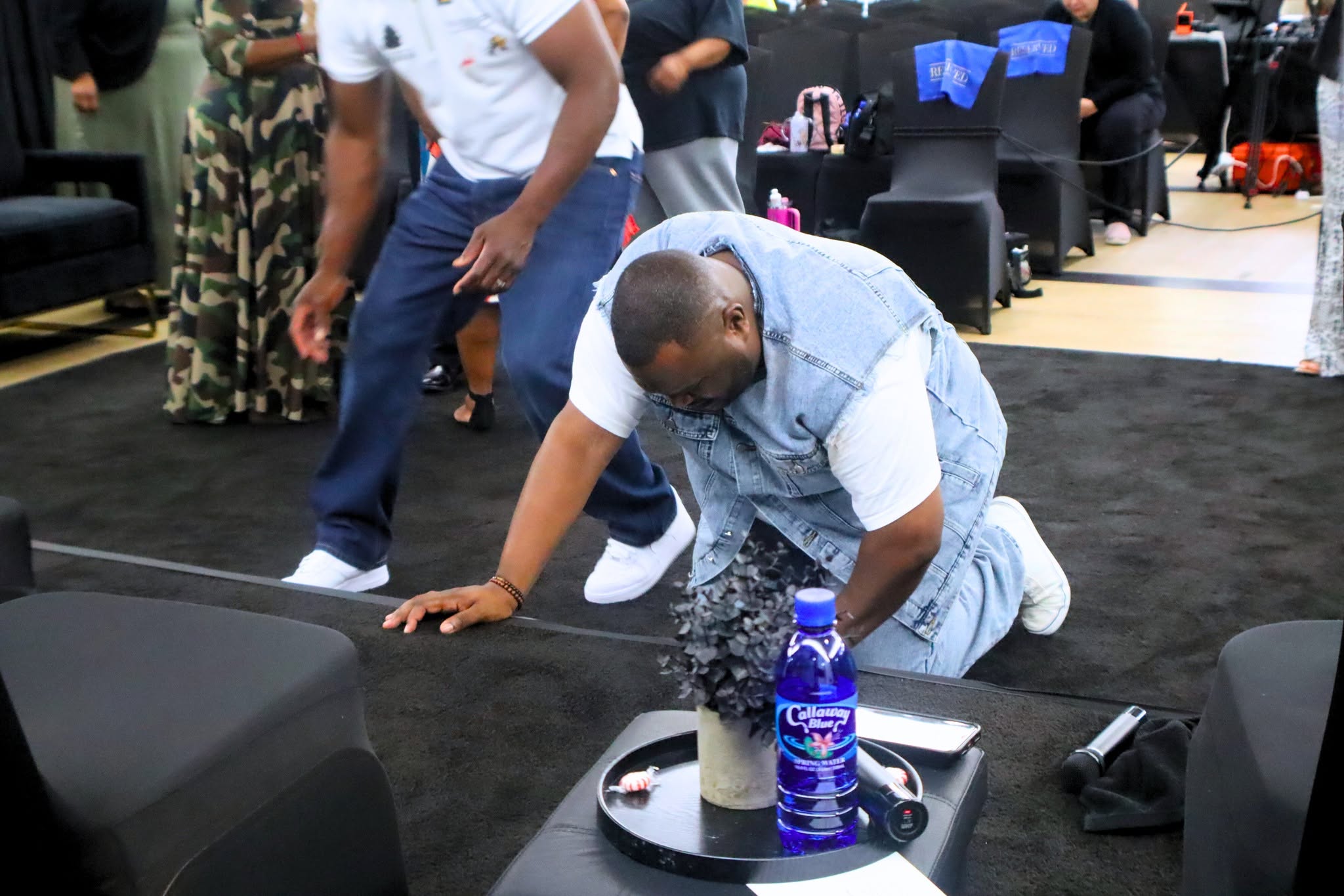 Person kneeling on black carpeted floor, reaching toward a tray with a water bottle and other items, surrounded by people standing and walking.