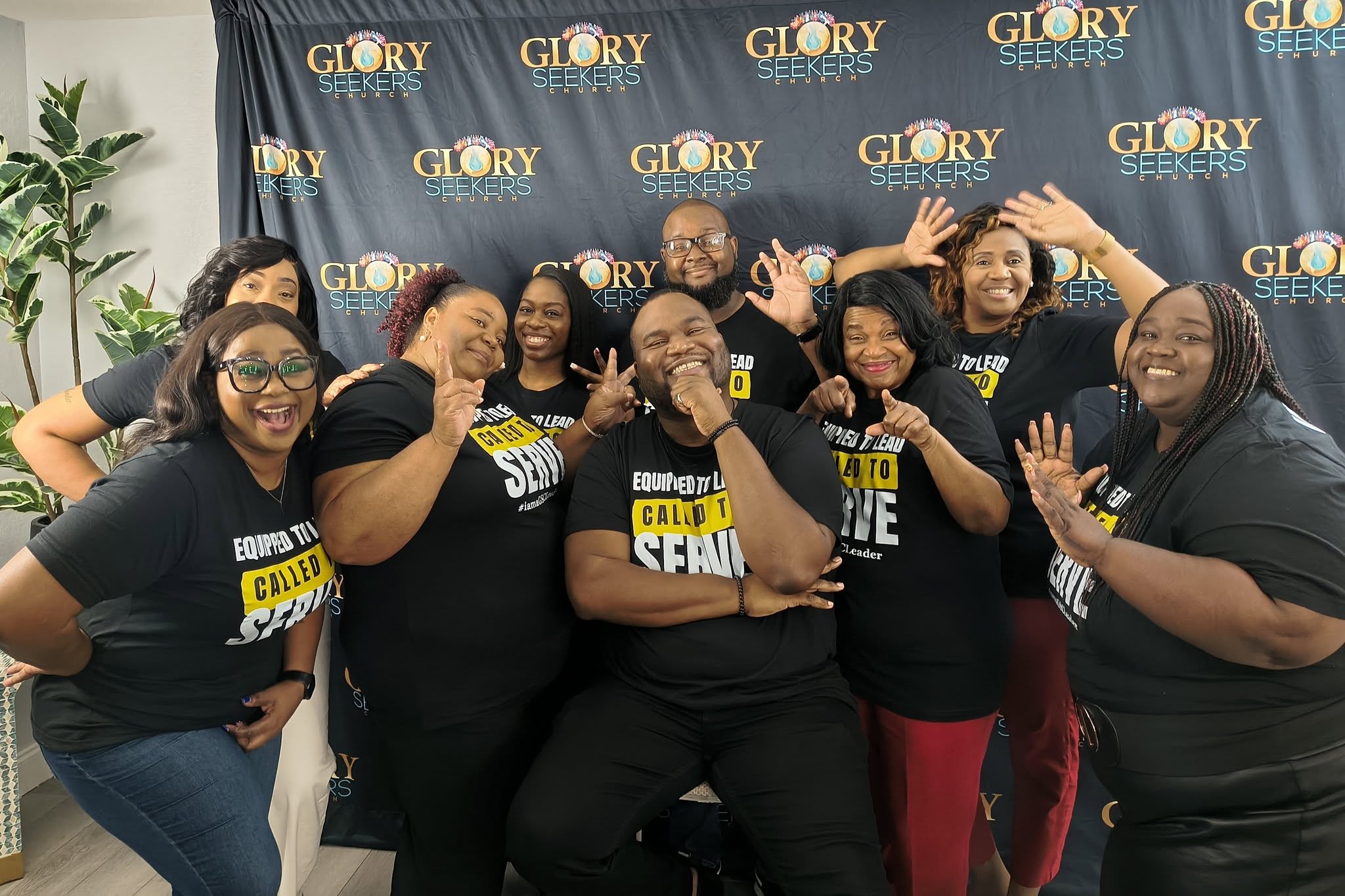 Group of nine people smiling and posing in front of a blue backdrop with 'Glory' logos, some making playful gestures.