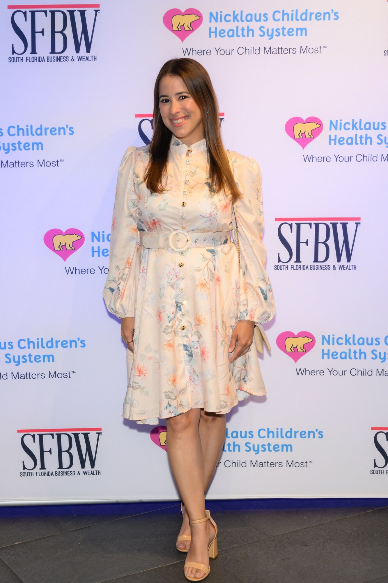 Young woman in a white floral dress standing in front of a backdrop with logos, smiling, with hands at sides.