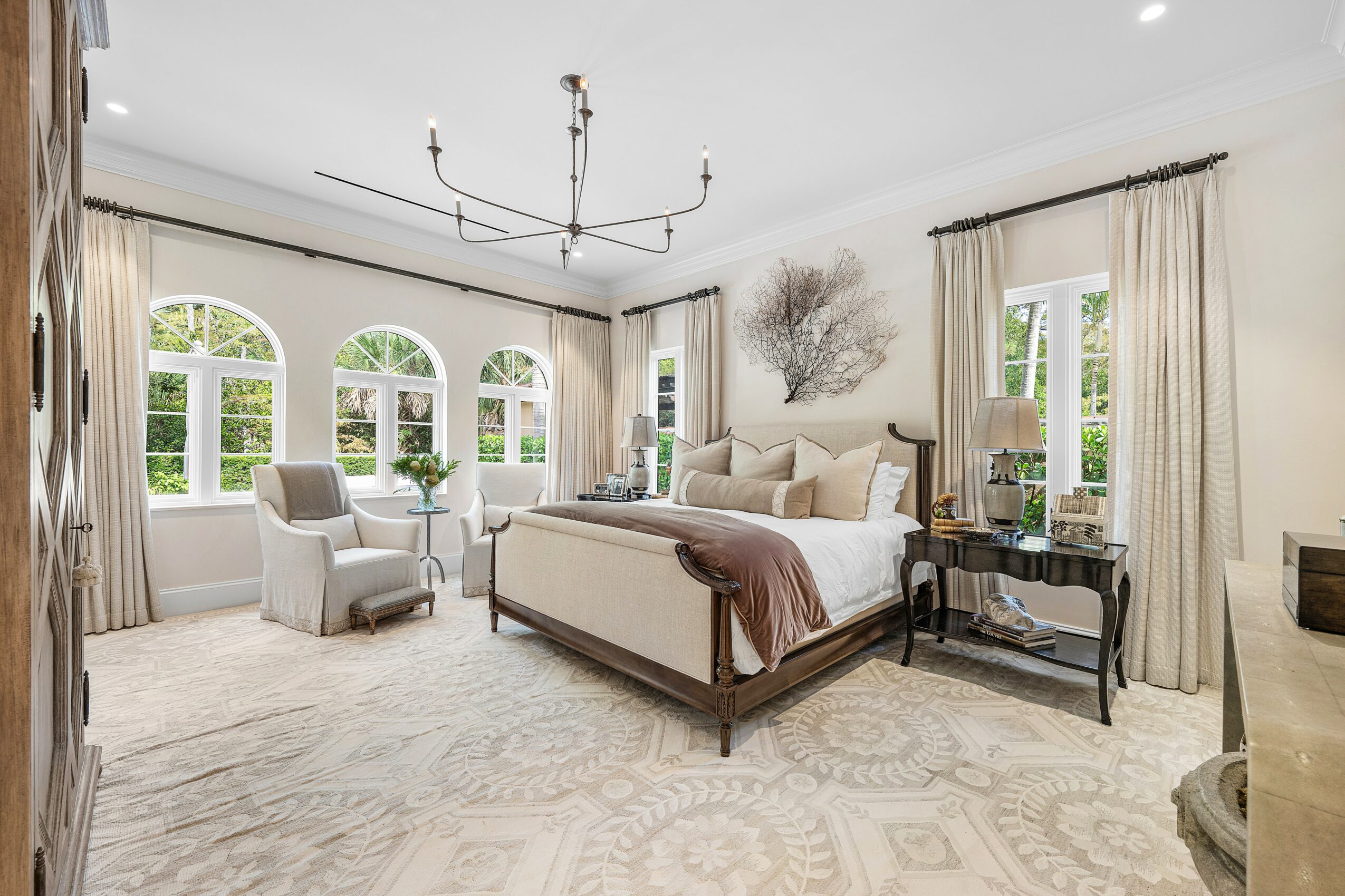 Elegant bedroom with large windows, a bed, seating area, and decorative plants, bright and airy with neutral tones.