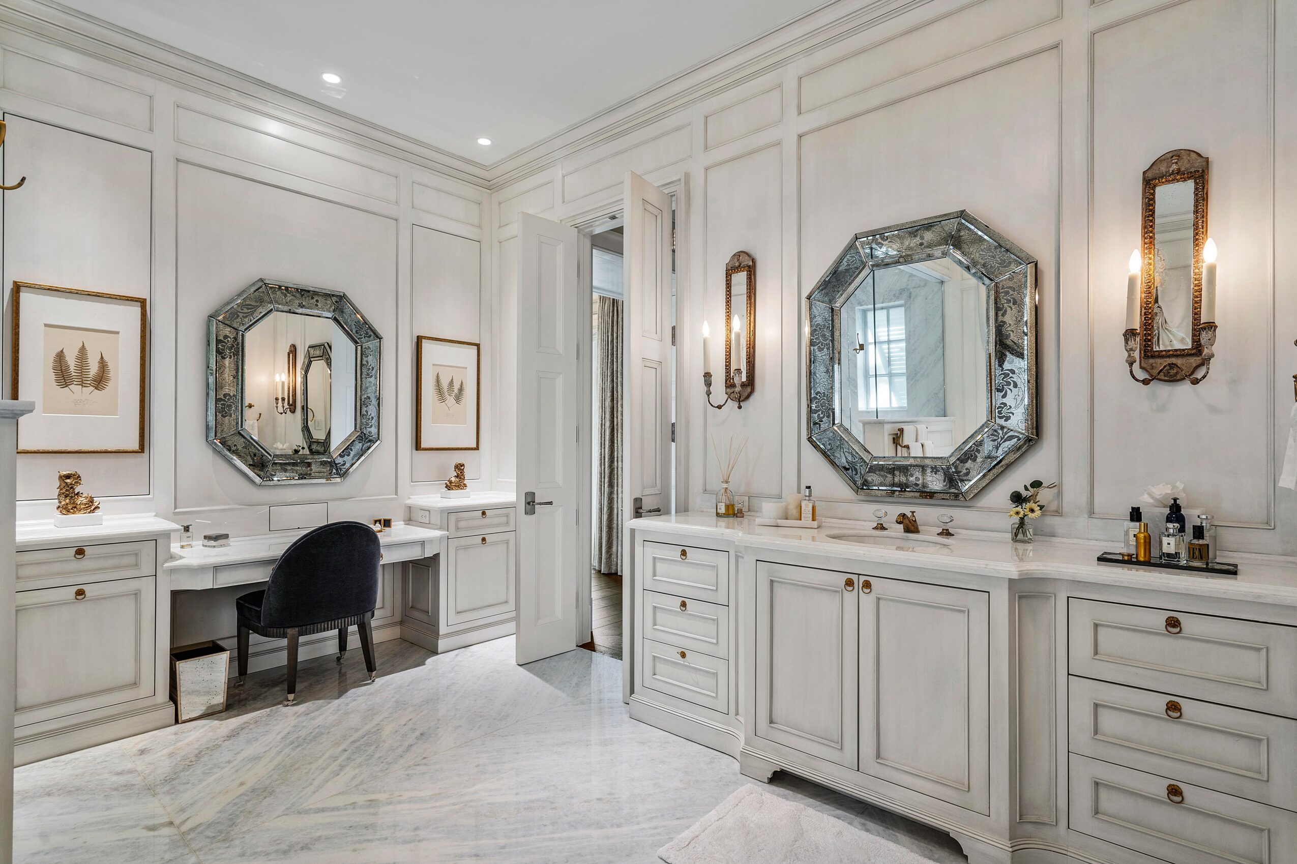 Elegant bathroom with white cabinetry, two mirrors, and a small vanity area with a black chair, decorated with wall sconces.