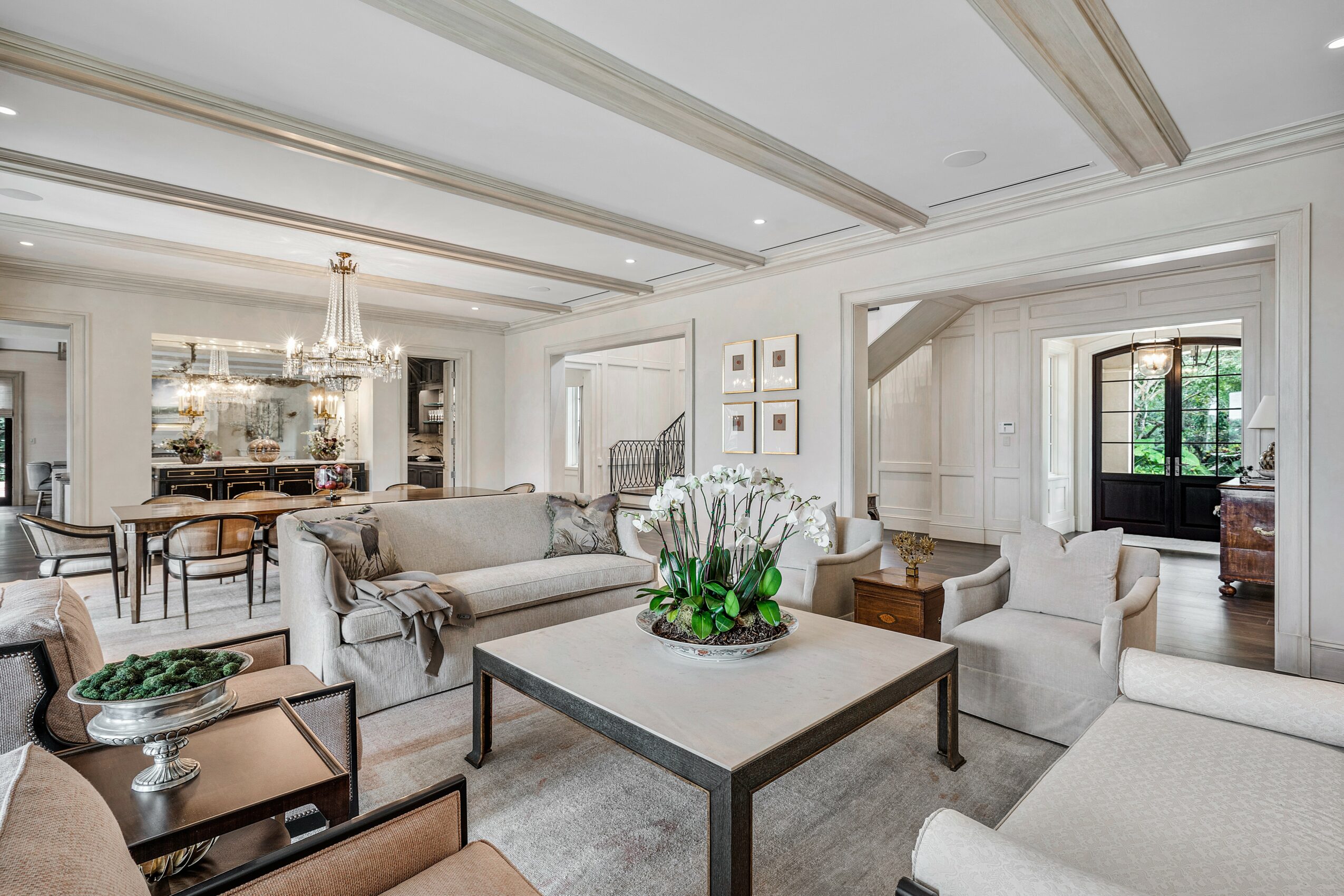 Living room with sofas, coffee table, and decorative items, connected to dining area with chandelier, bright and elegant interior.