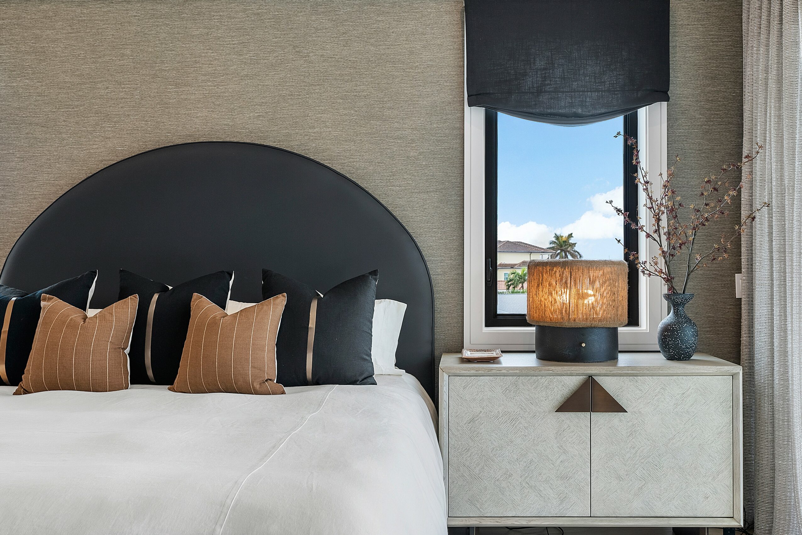 Bedroom with a bed, black and brown pillows, a nightstand with a lamp, and a window with curtains and a view outside.