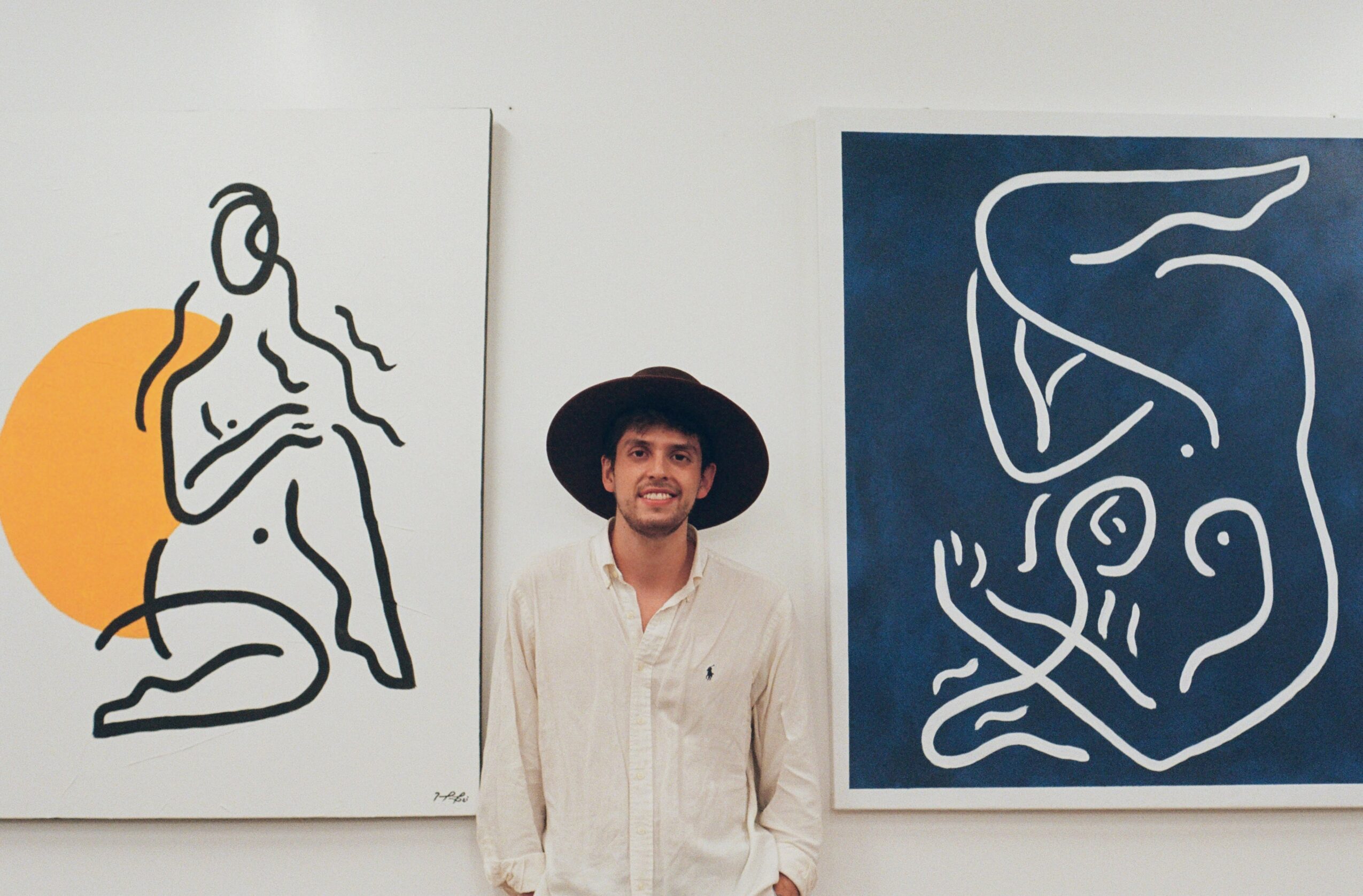 Person with a wide-brimmed hat standing between two abstract line art paintings on a white wall.