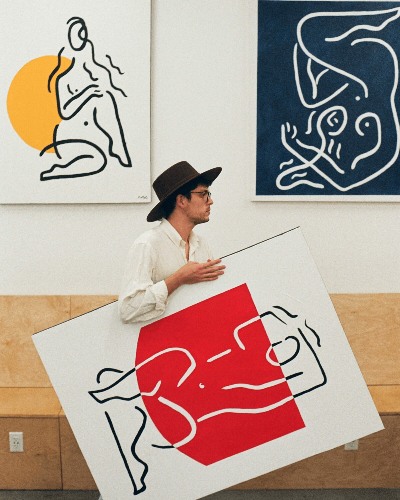 Person holding a large artwork with a red background and black line drawing, wearing a hat and glasses, in an art gallery.