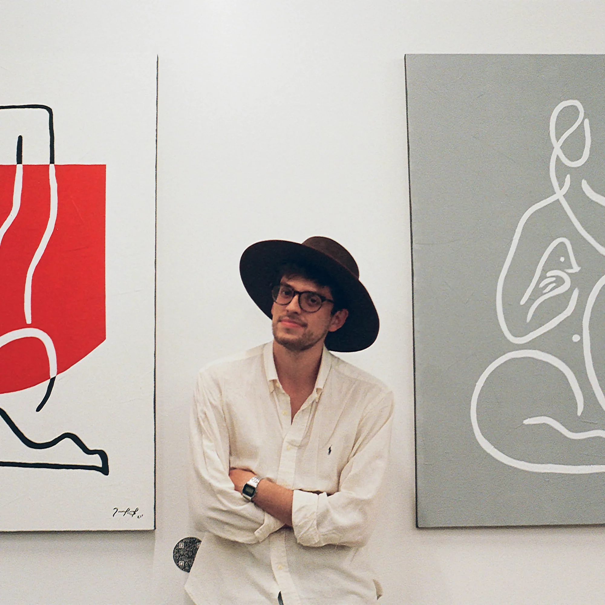 Young man with glasses and a wide-brimmed hat, wearing a light-colored shirt, standing with arms crossed in front of abstract art.