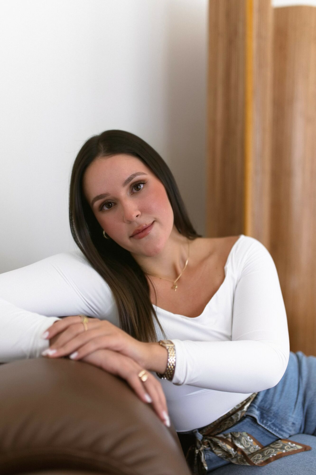 Woman with long dark hair lying on a bed, wearing a white top and jeans, looking at the camera.
