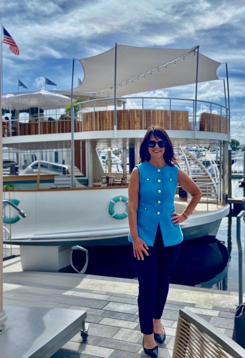 Woman in sunglasses and blue sleeveless top standing on dock in front of a boat at marina under partly cloudy sky.