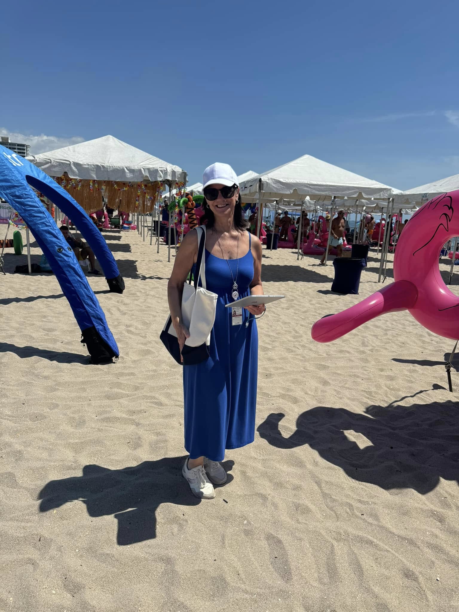 Woman in blue dress and white hat standing on sandy beach with tents and inflatable structures in background.