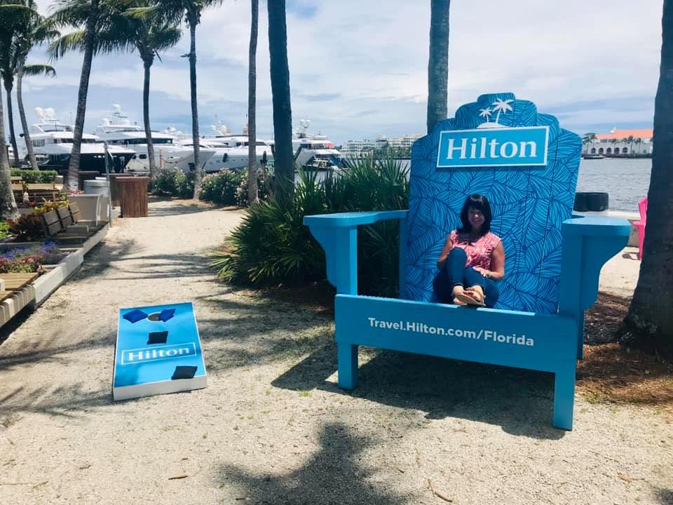 Person sitting on a large blue Hilton chair outdoors near palm trees and water, with a Hilton sign and a small blue stand nearby.