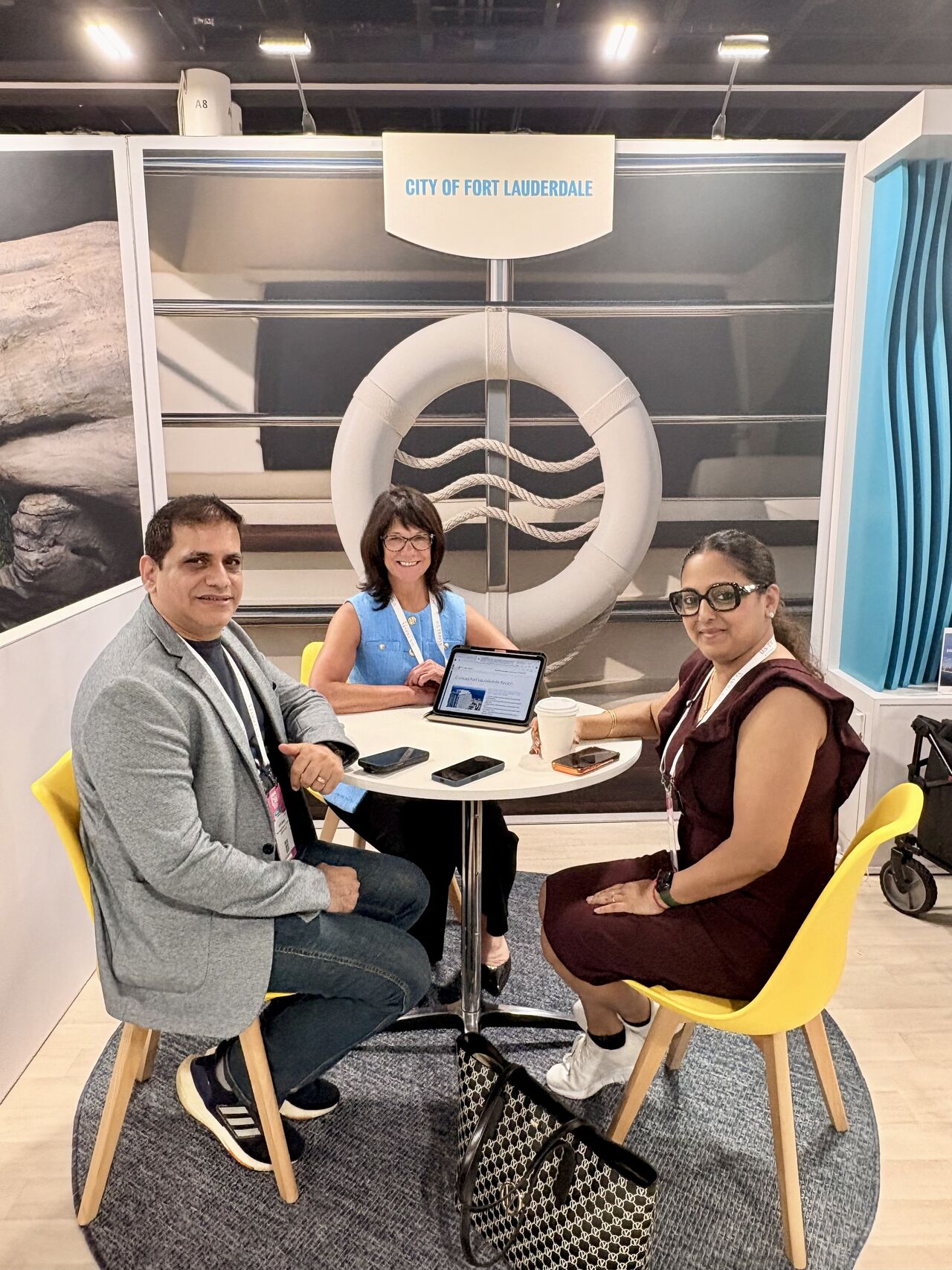 Three people sitting around a table with a laptop and tablet, in front of a backdrop with a large lifebuoy and a sign reading 'City of Fort Lauderdale'.