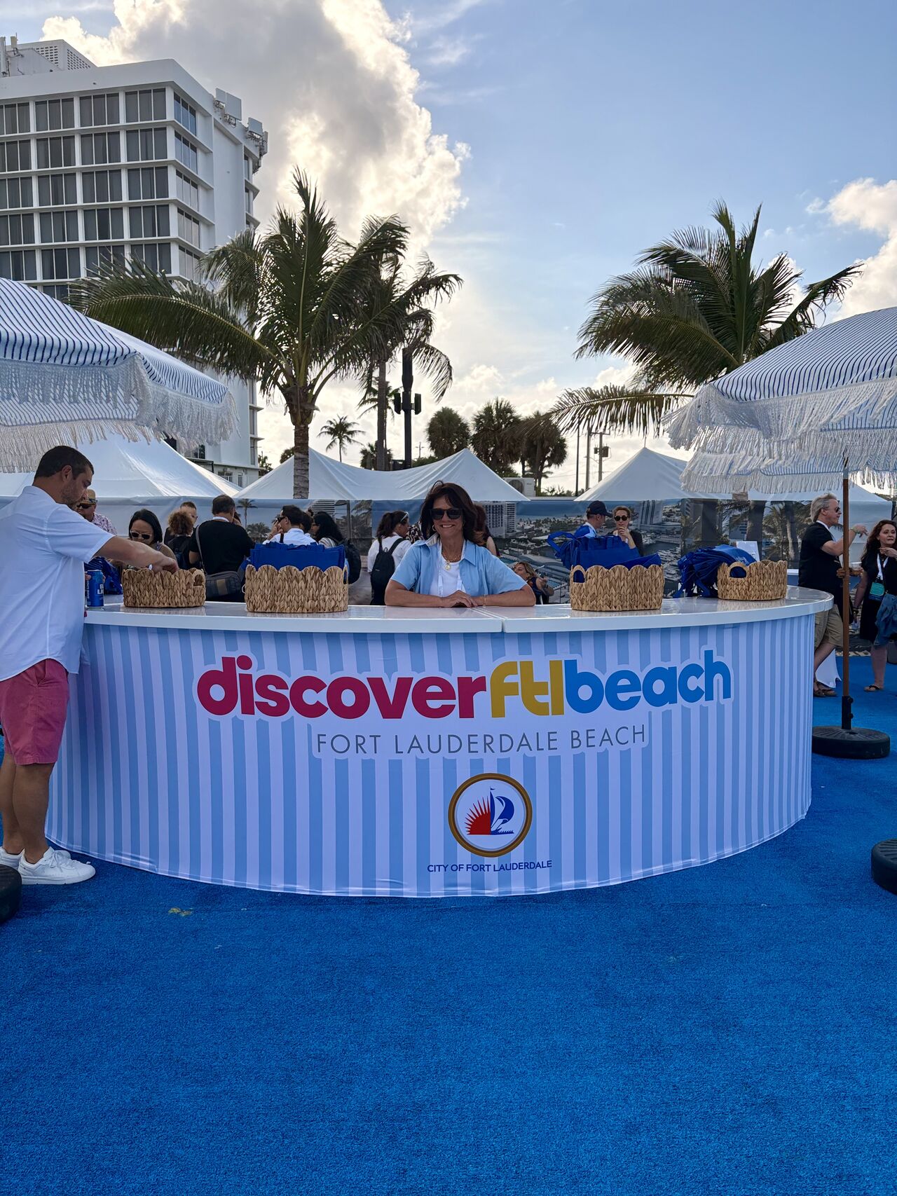People gather around a Discover FL Beach booth at Fort Lauderdale Beach with palm trees and buildings in the background.