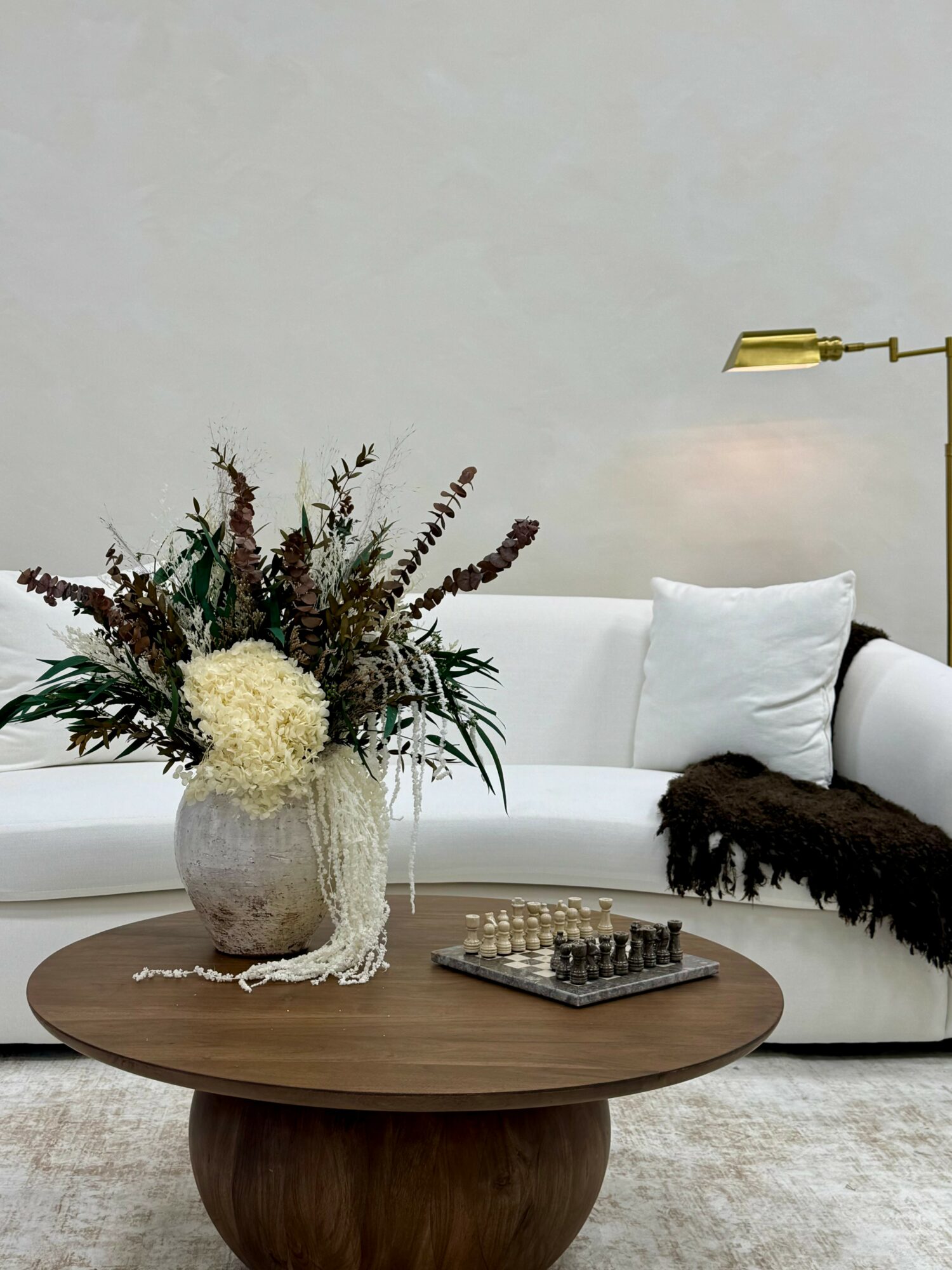 Living room with a white sofa, a wooden coffee table with a large flower vase and a chessboard, and a floor lamp.