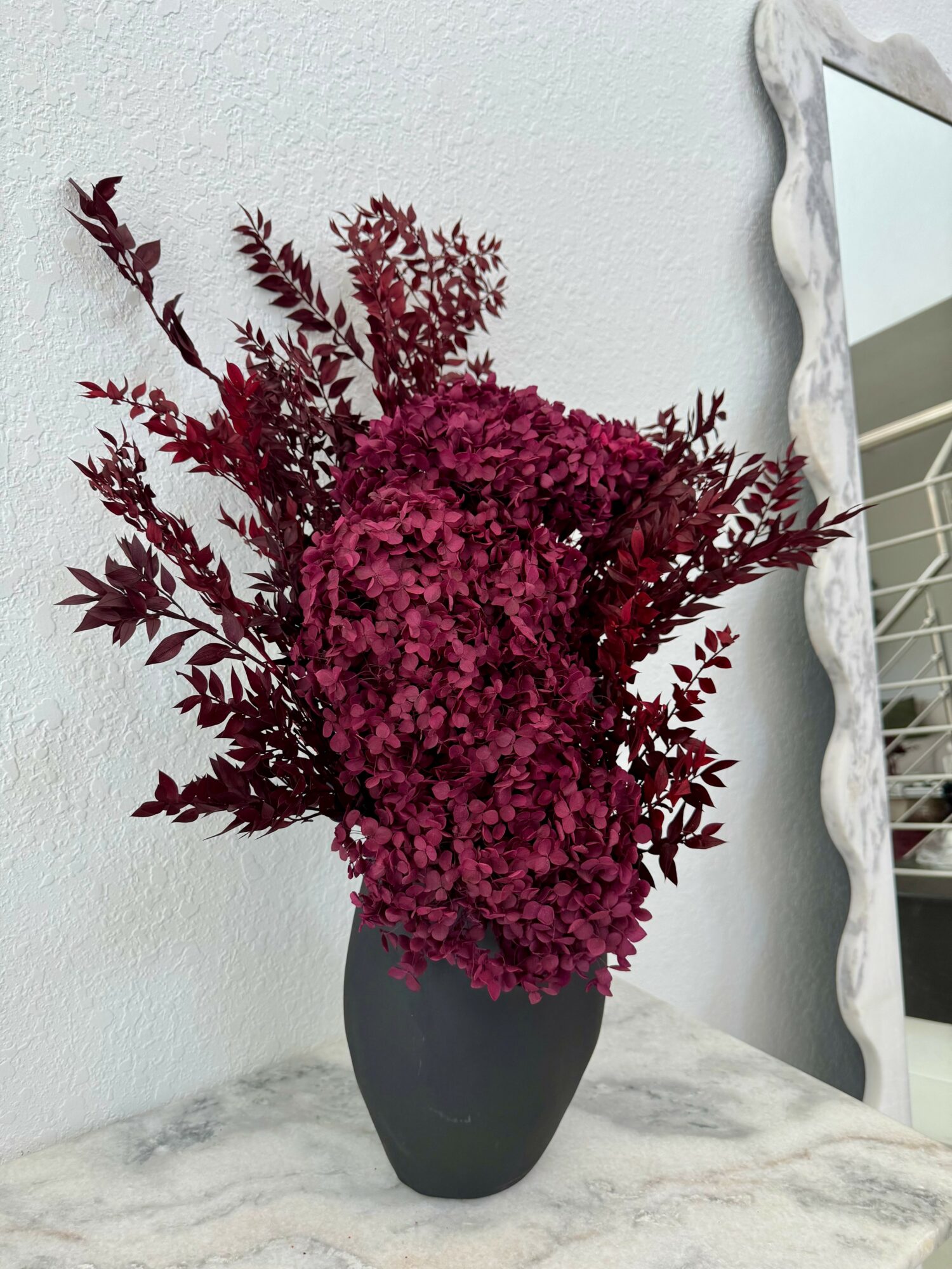 Dark pink and burgundy flowers in a black vase on a marble surface, with a mirror and white wall in background.