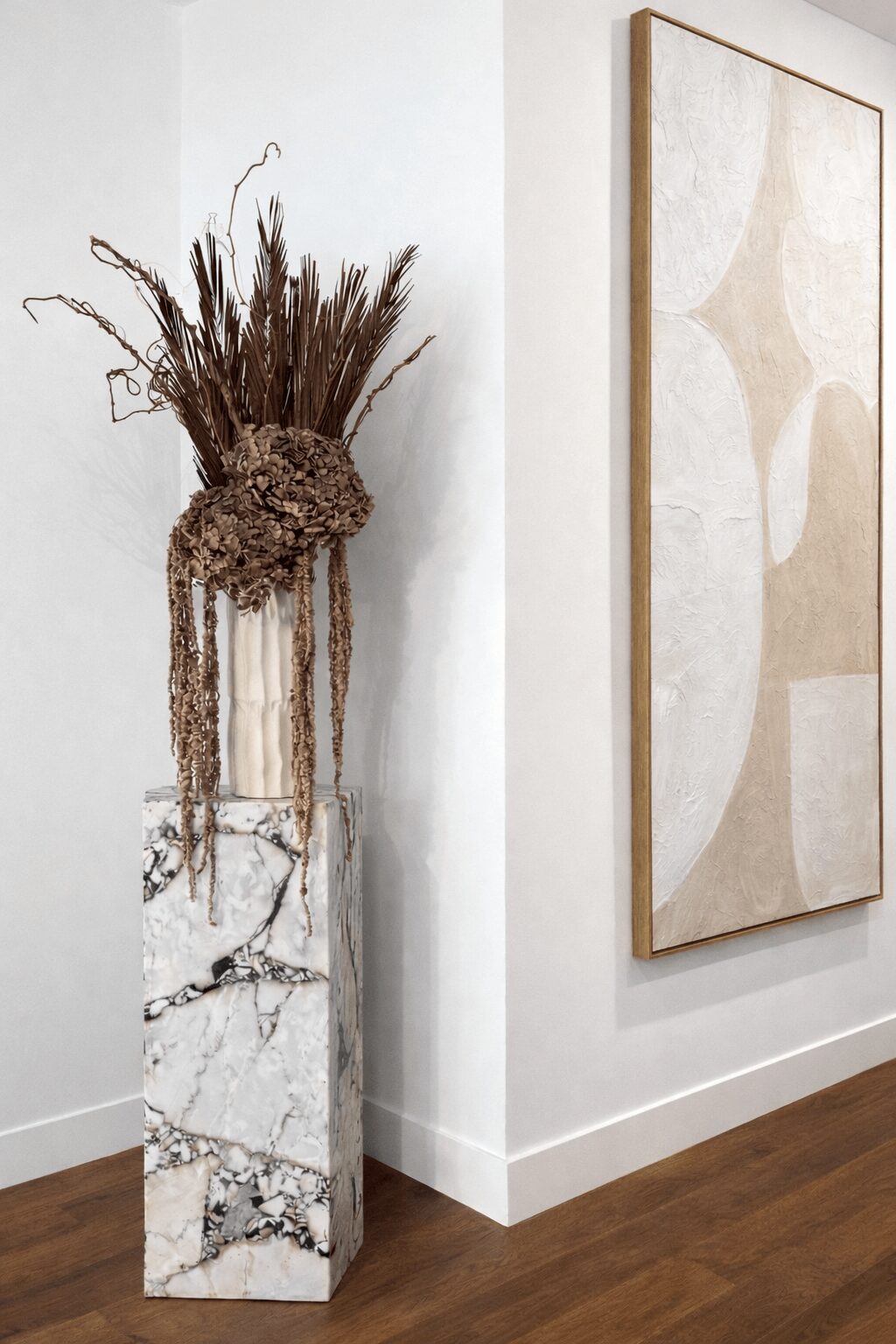 Tall marble vase with dried flowers and branches next to a large abstract artwork on a white wall.