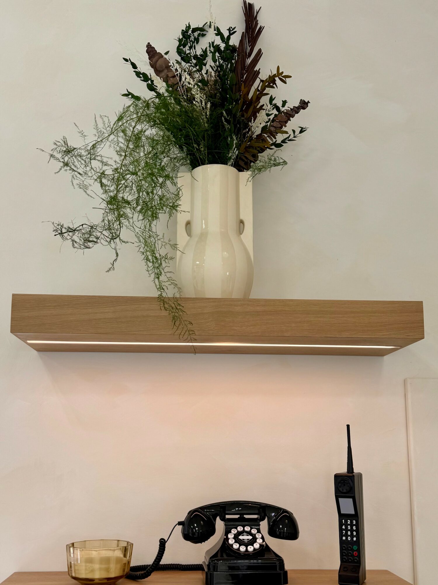 Vase with dried flowers on a wall shelf, with a vintage rotary phone and a cordless phone below.