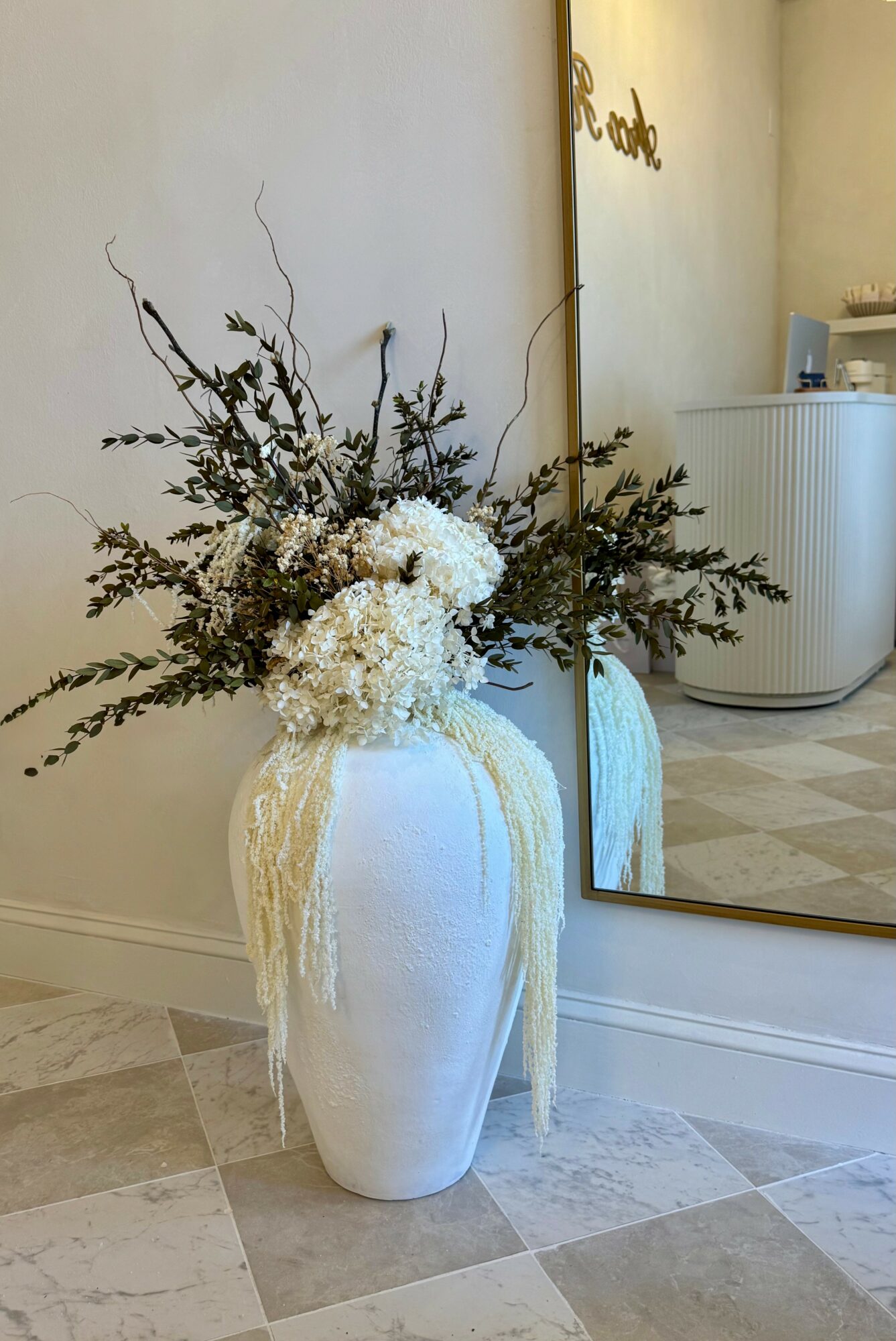 White vase with dried flowers and branches, placed on a tiled floor near a mirror, with a wall and a sign in the background.