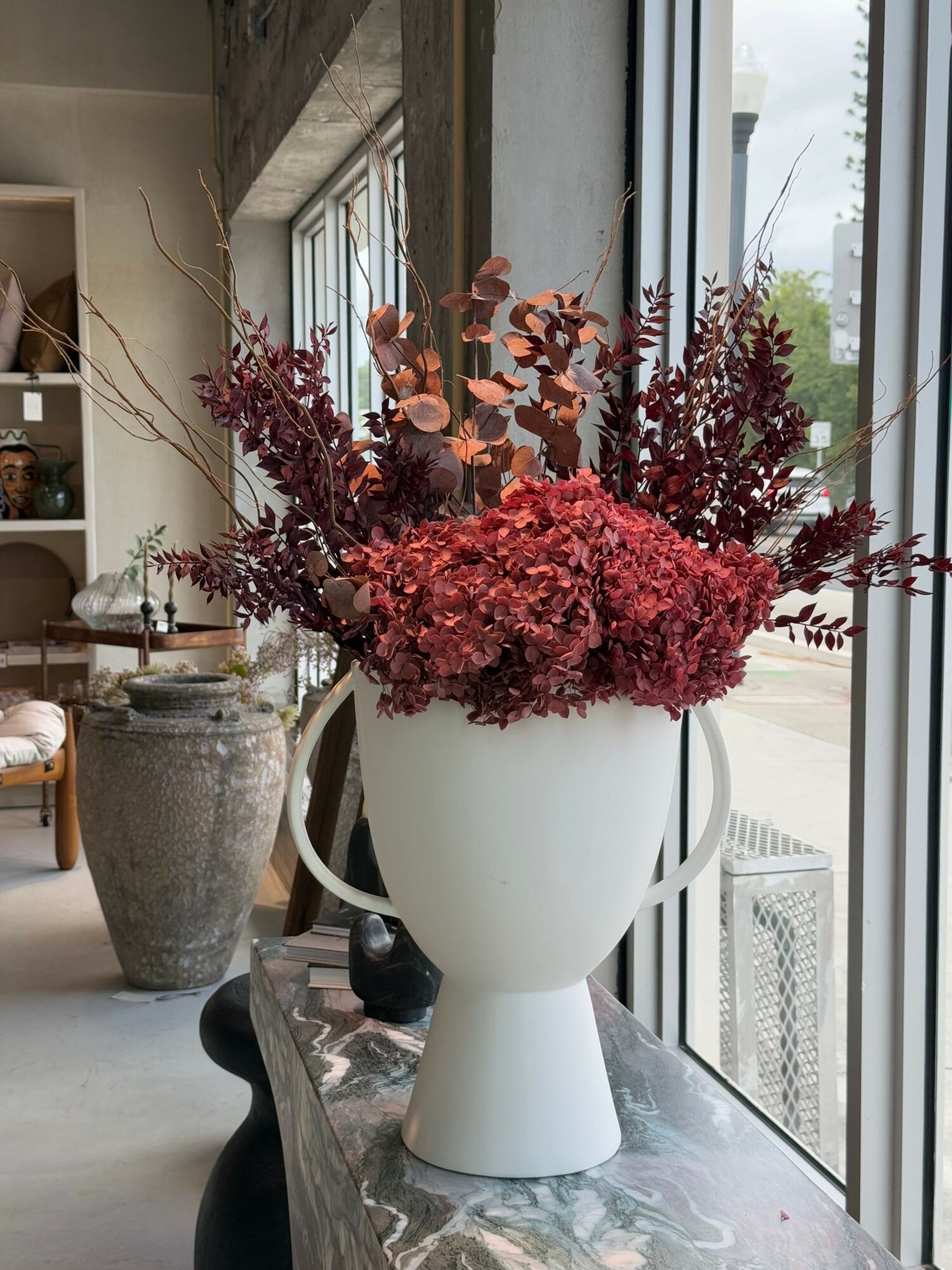 White vase with pink and red flowers on a reflective surface near a window, with a potted plant in the background.