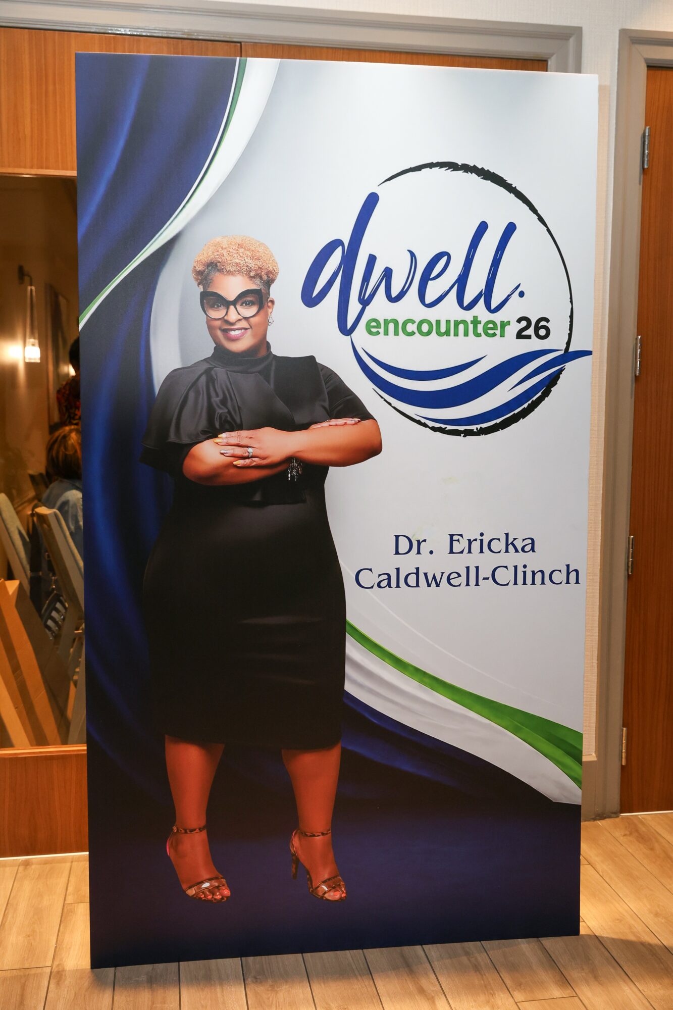 Standing woman with short blonde hair and glasses, wearing a black dress, in front of a sign with text and logo.