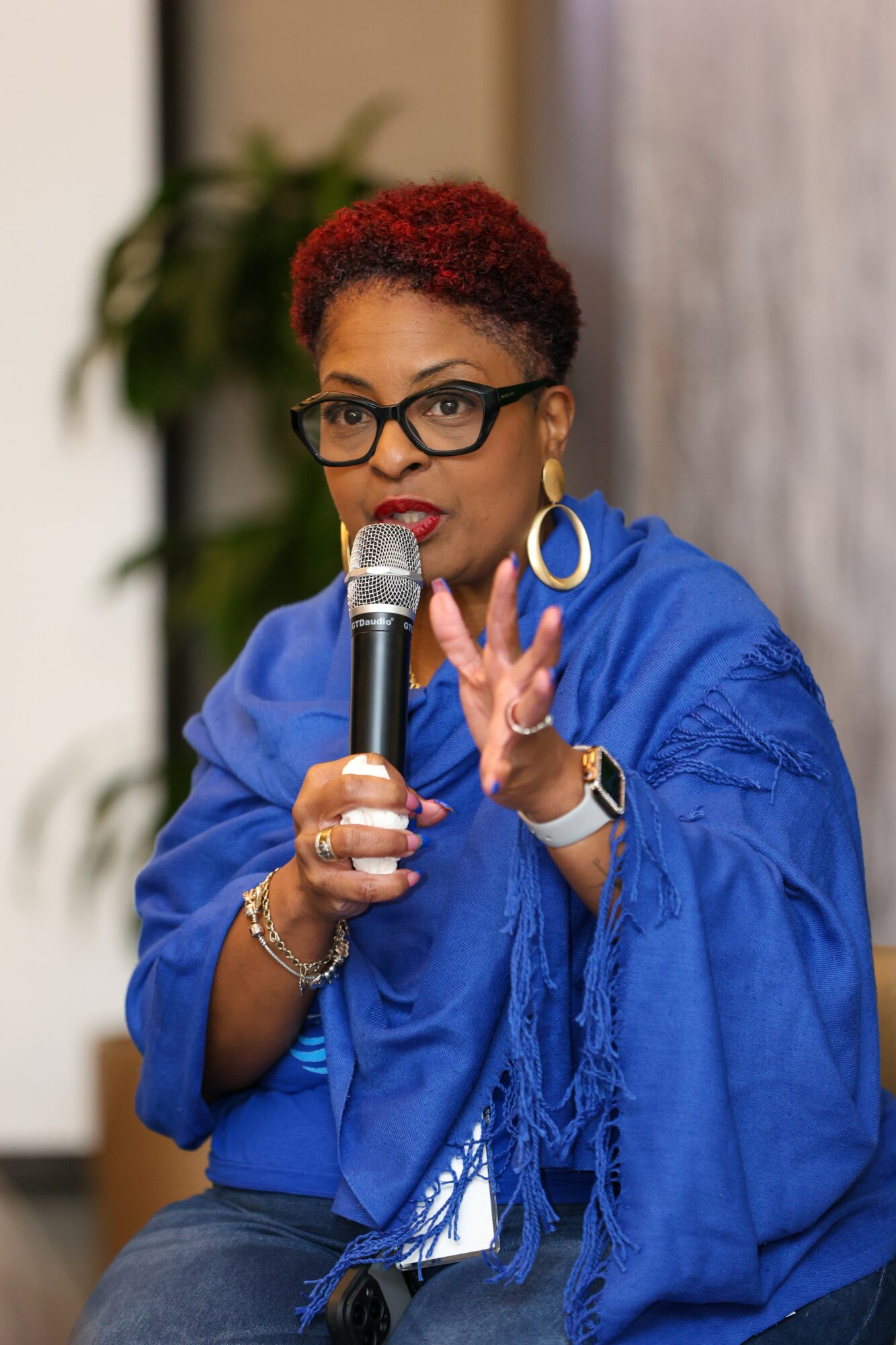 Woman with short red hair, glasses, and large earrings speaking into a microphone, gesturing with her hand, wearing a blue outfit.